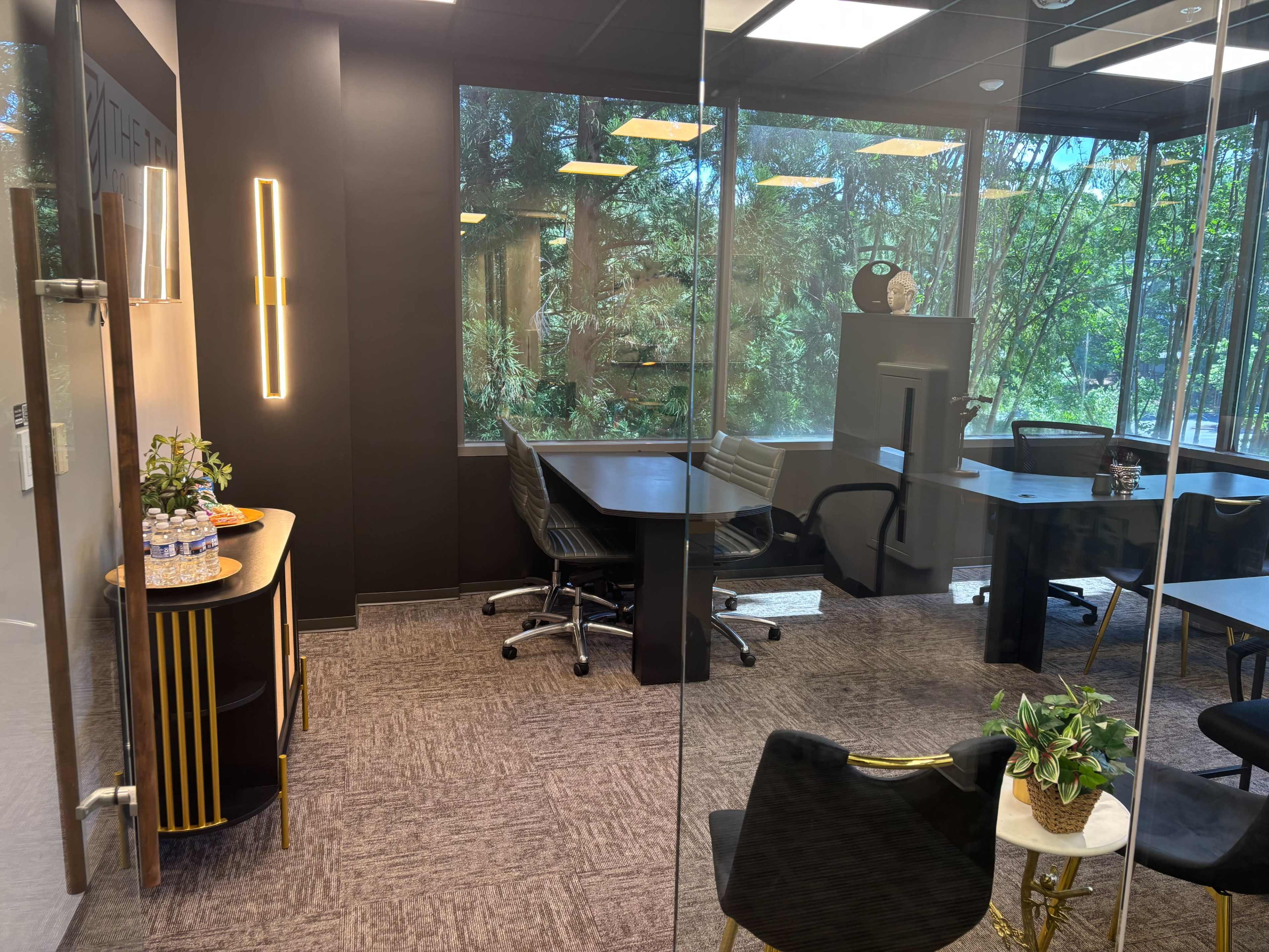 The image shows a modern office space with glass walls, several black office chairs, a round table, and a view of greenery outside.