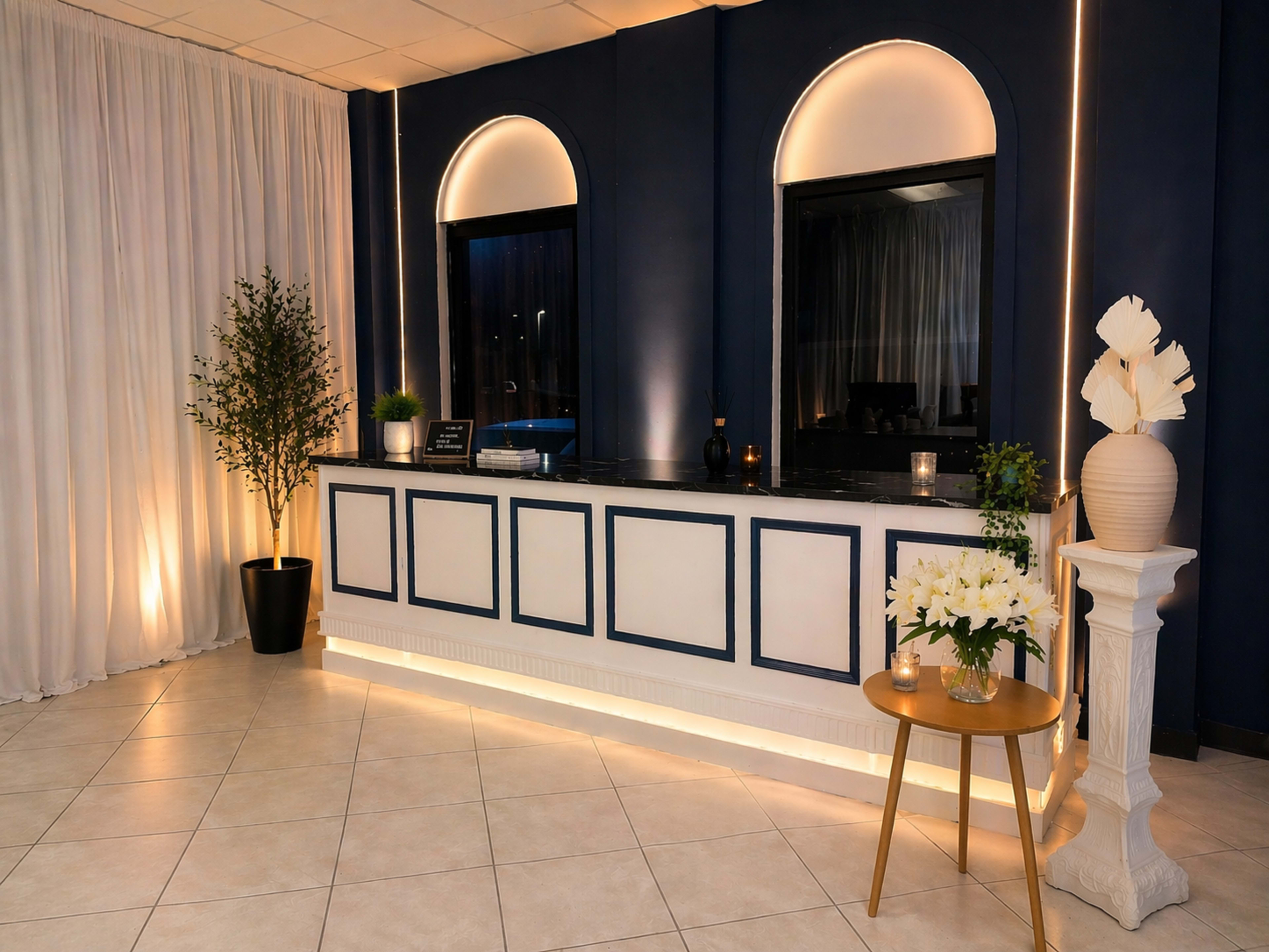 The image shows a modern reception area with a dark blue wall, a white counter with arched windows, and decorative plants and flowers placed around the space.