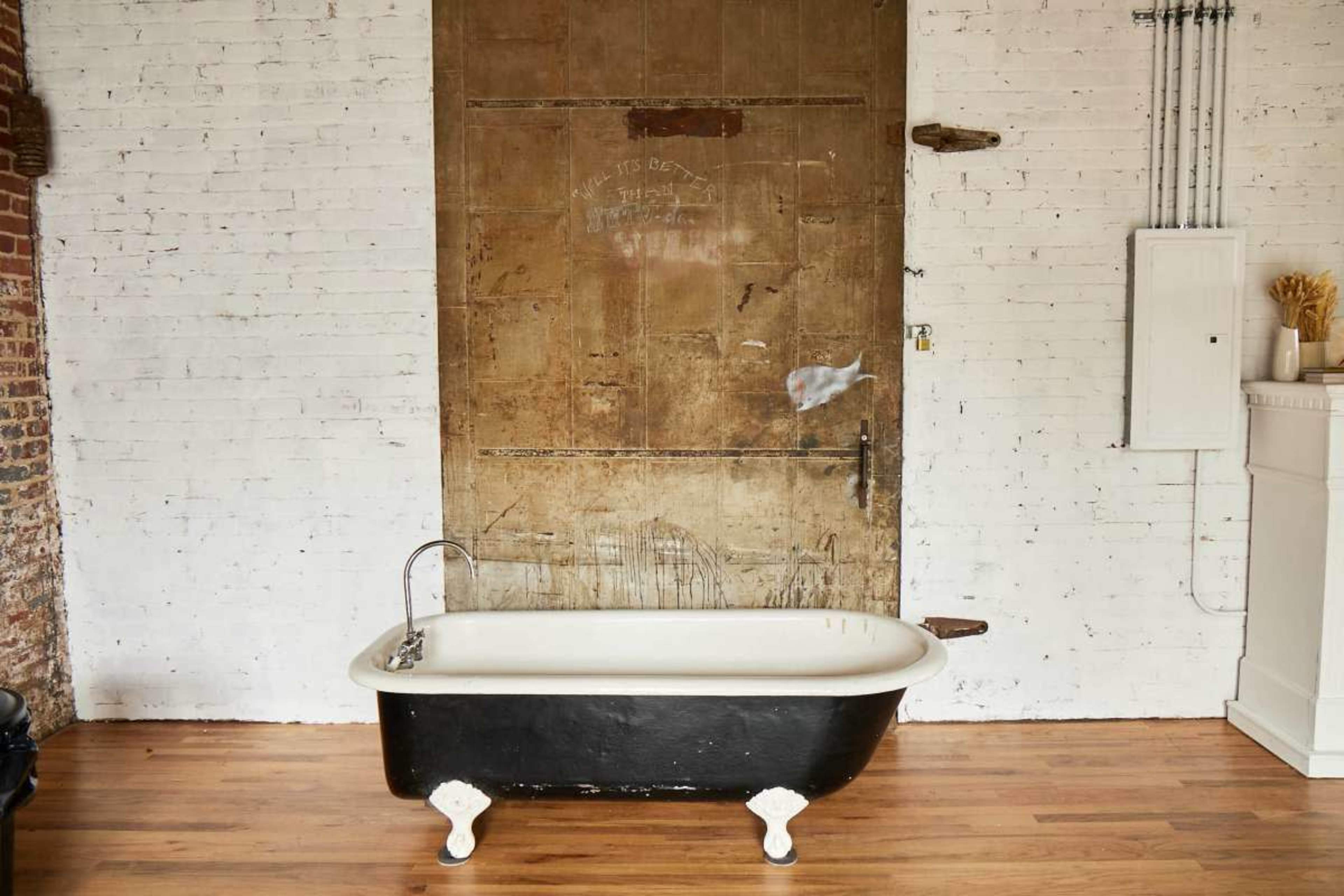 A vintage black and white freestanding bathtub is positioned in front of a weathered wooden door against a white brick wall.