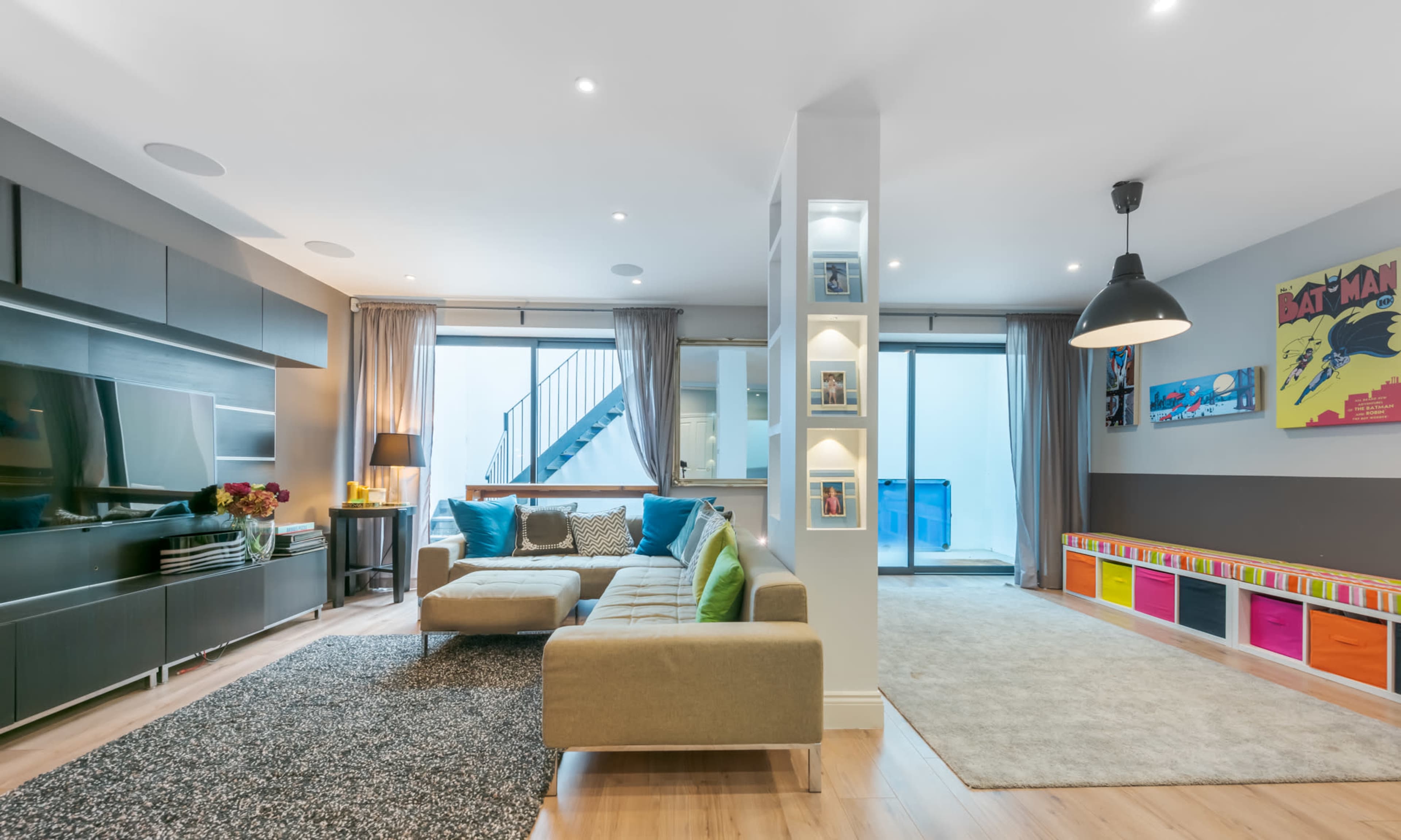 The image shows a modern living room with a sectional sofa, a television unit, and colorful storage bins along one wall, adjacent to a staircase and a large window.