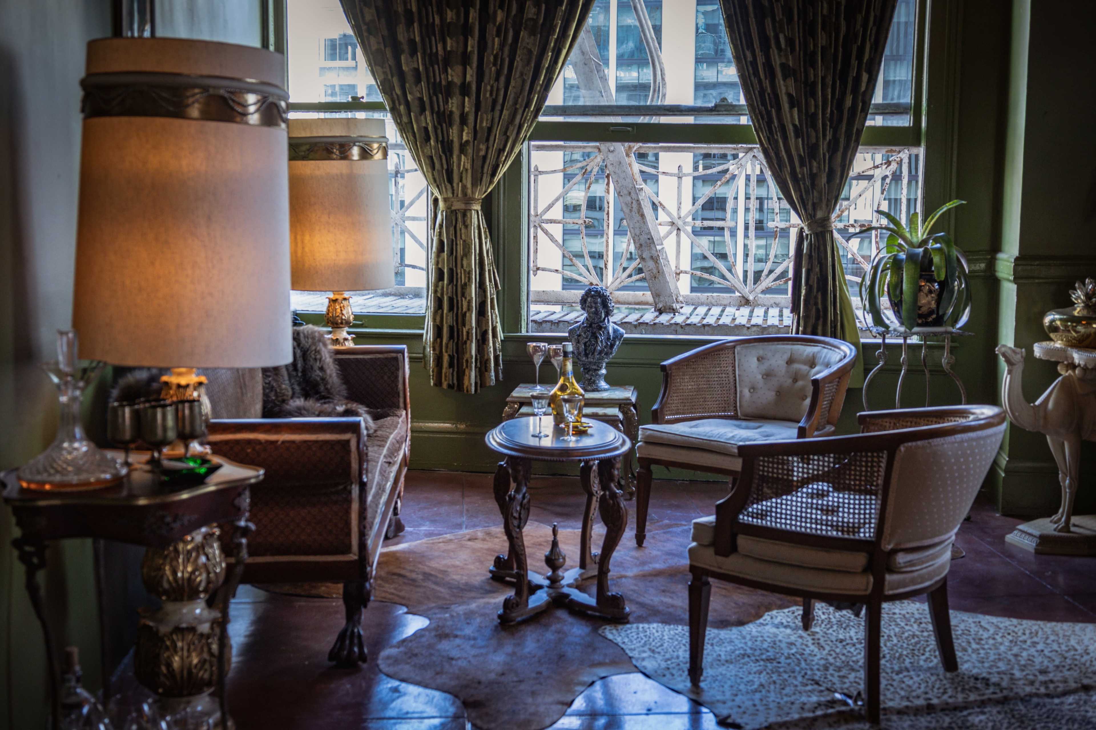 A cozy sitting area features vintage furniture, including two chairs, a table, and a decorative lamp, with sunlight streaming through a large window.