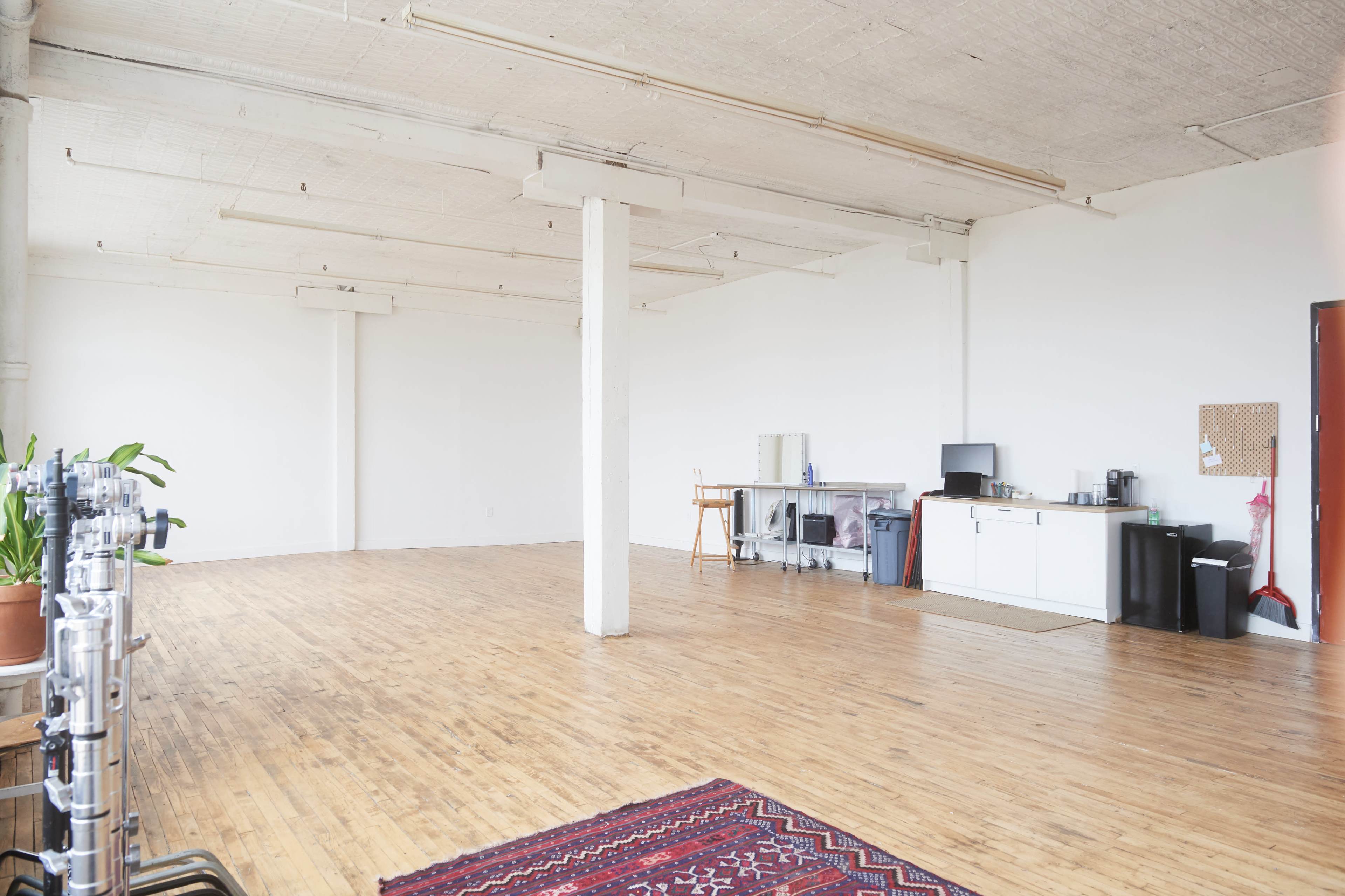 A spacious, empty room with wooden flooring, white walls, and a small kitchenette in the corner.