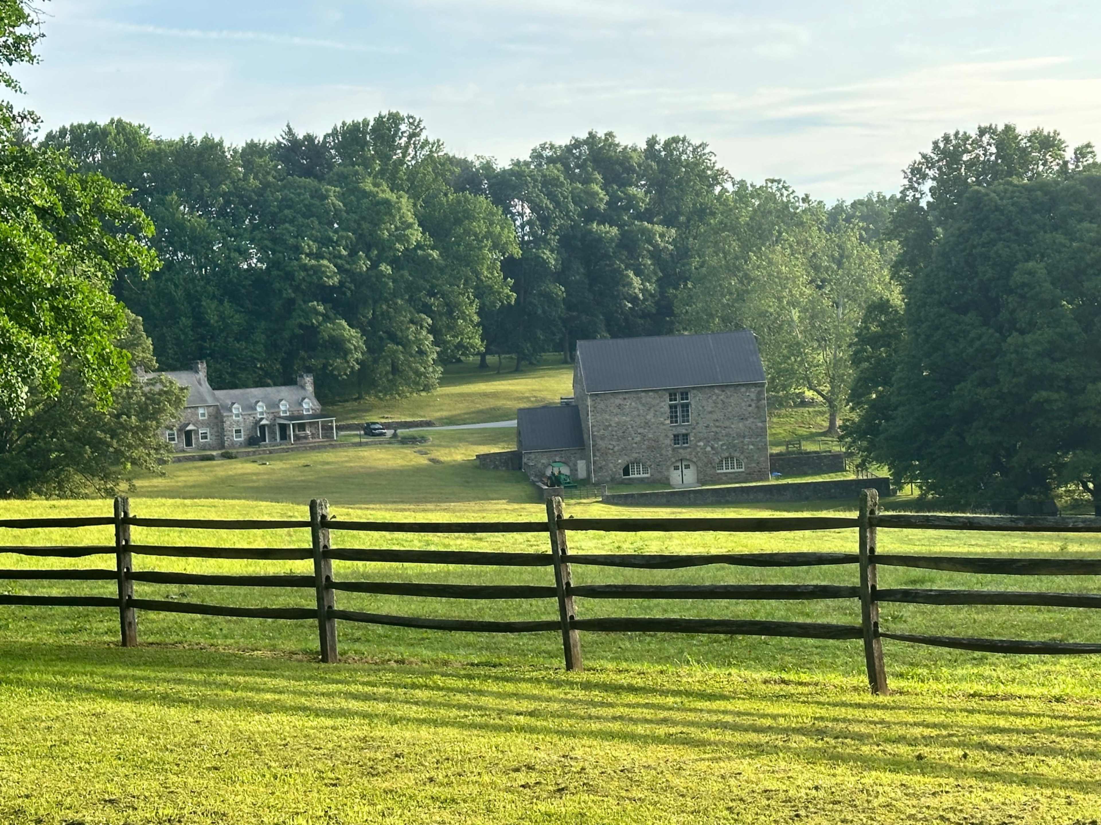 A stone house and a separate building set against a backdrop of lush green trees and a wooden fence.