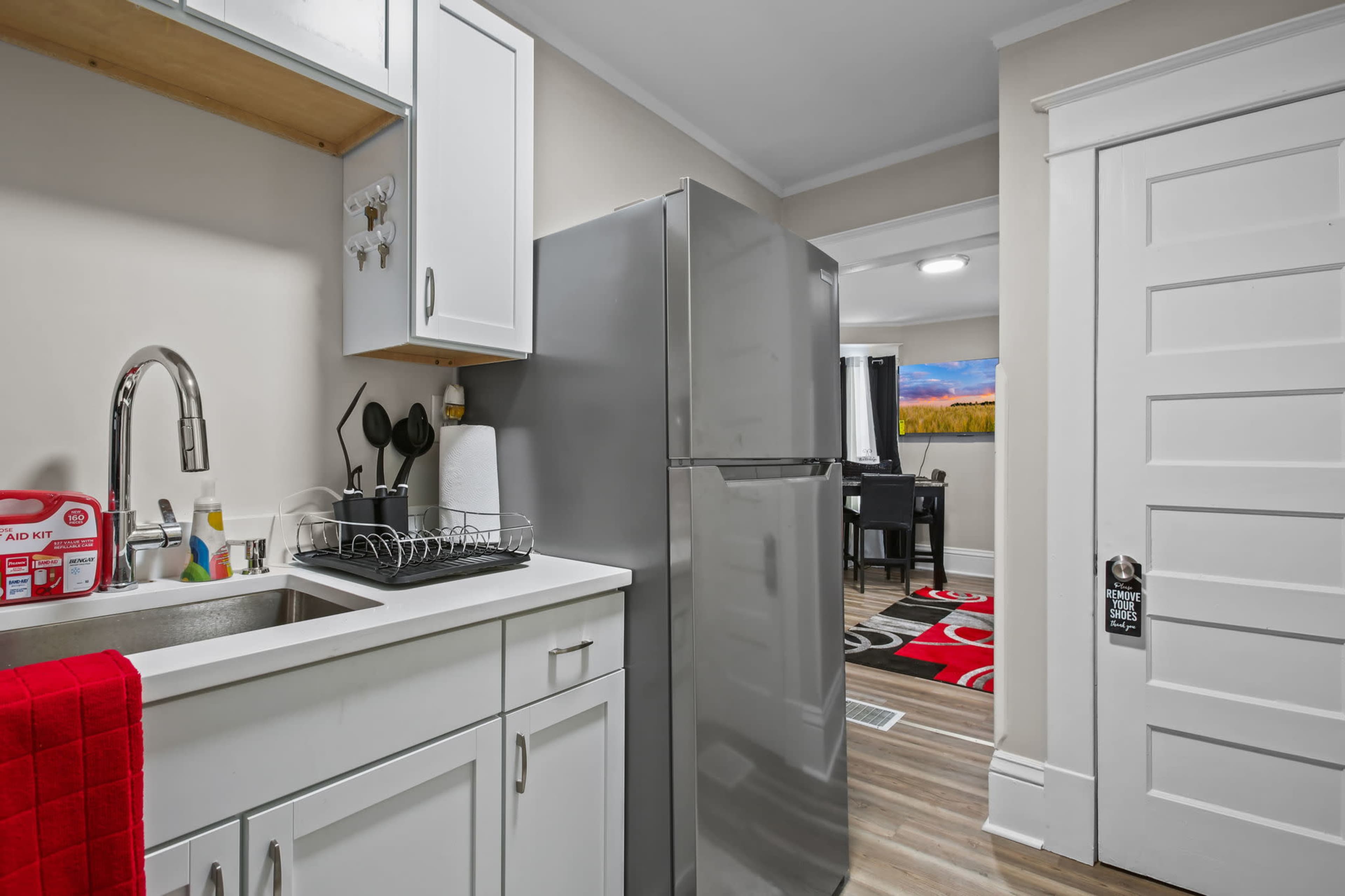 A compact kitchen with a stainless steel refrigerator, a sink, and white cabinetry, alongside a view of a dining area featuring a patterned rug.
