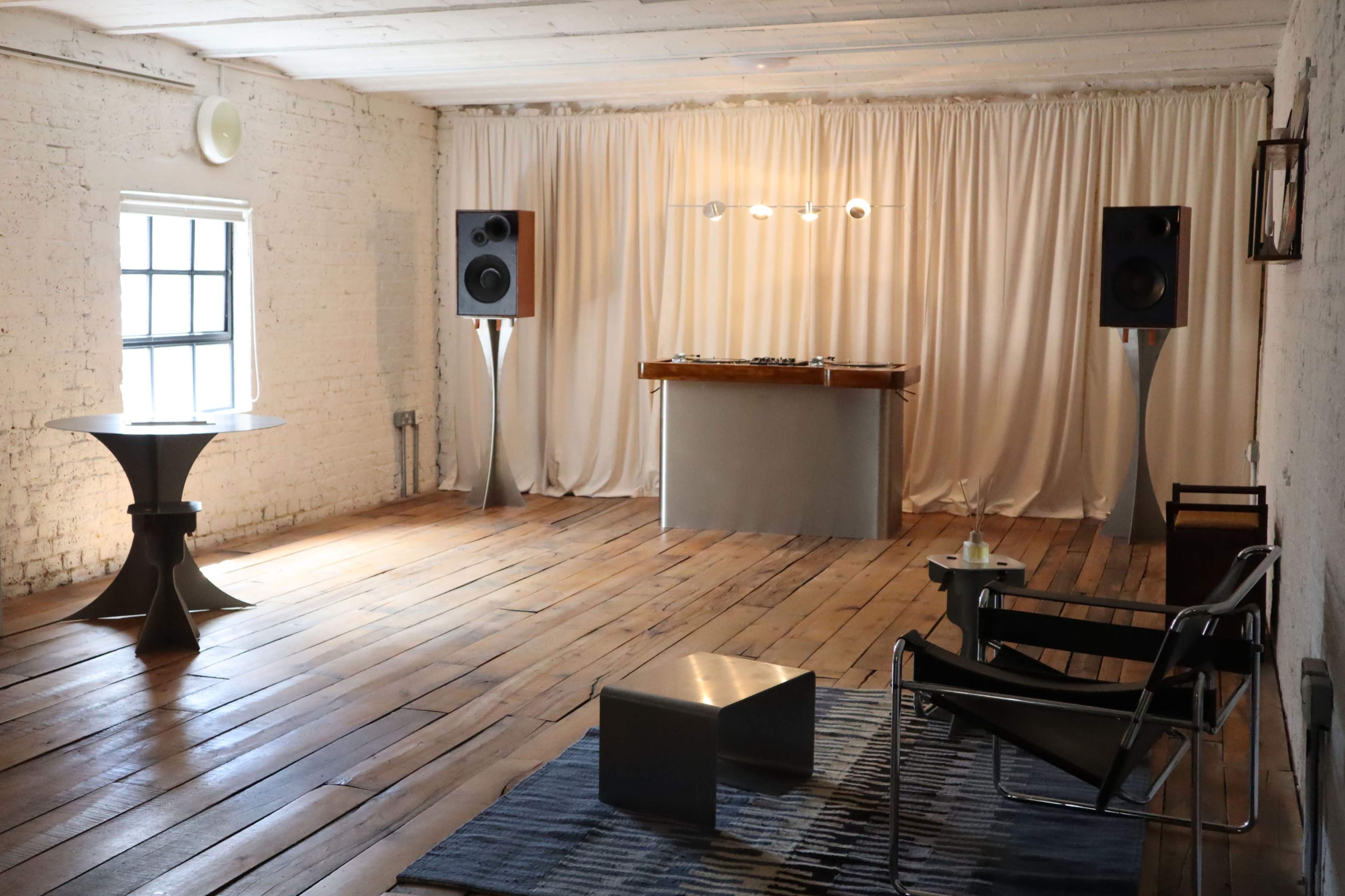 The image shows a minimalist room featuring a wooden floor, a DJ booth with speakers, a small table, and a chair.