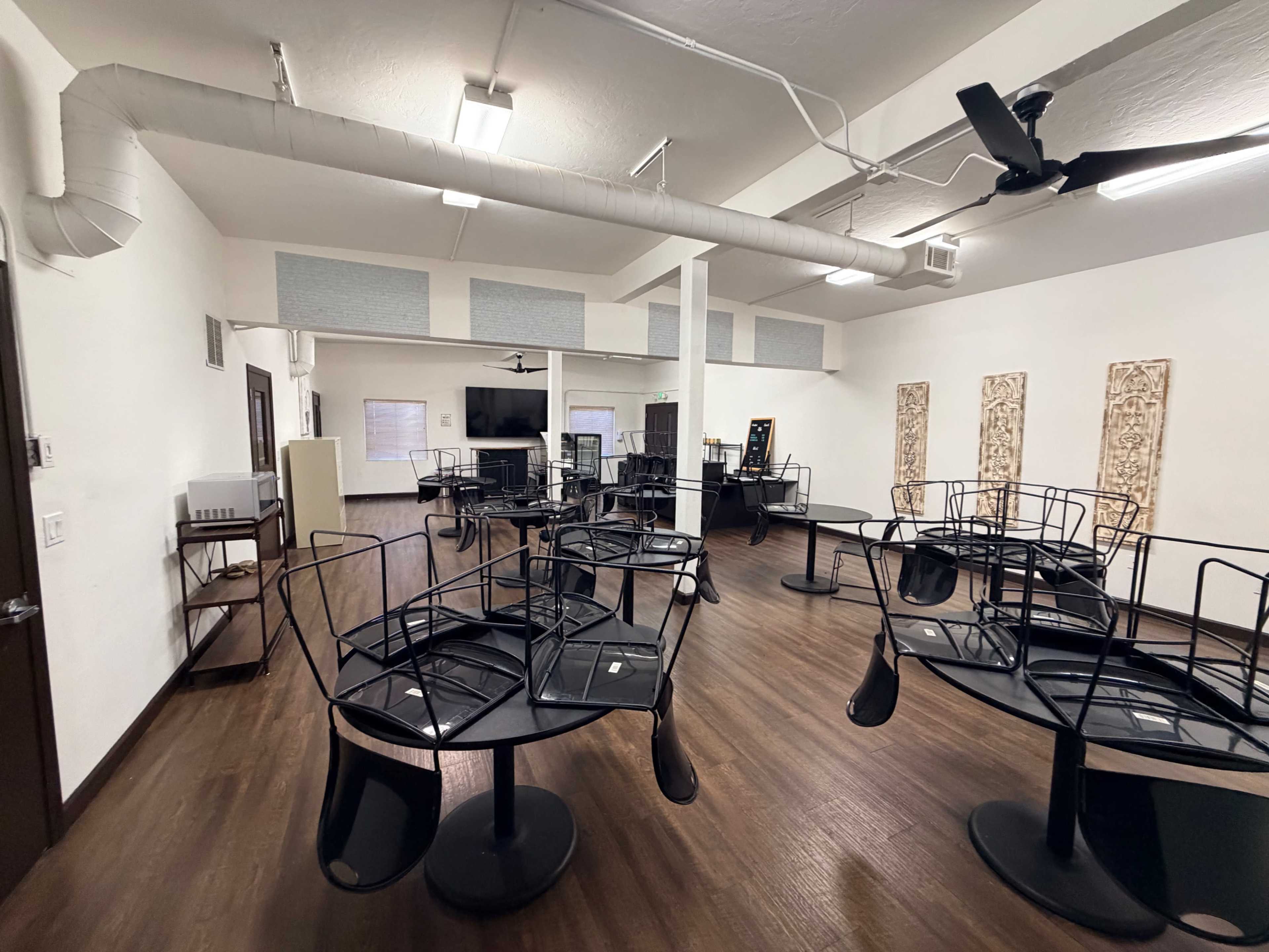 The image shows an empty room with several tables turned upside down on their stands, with a few chairs stacked beside them, and a large flat screen visible on the wall.