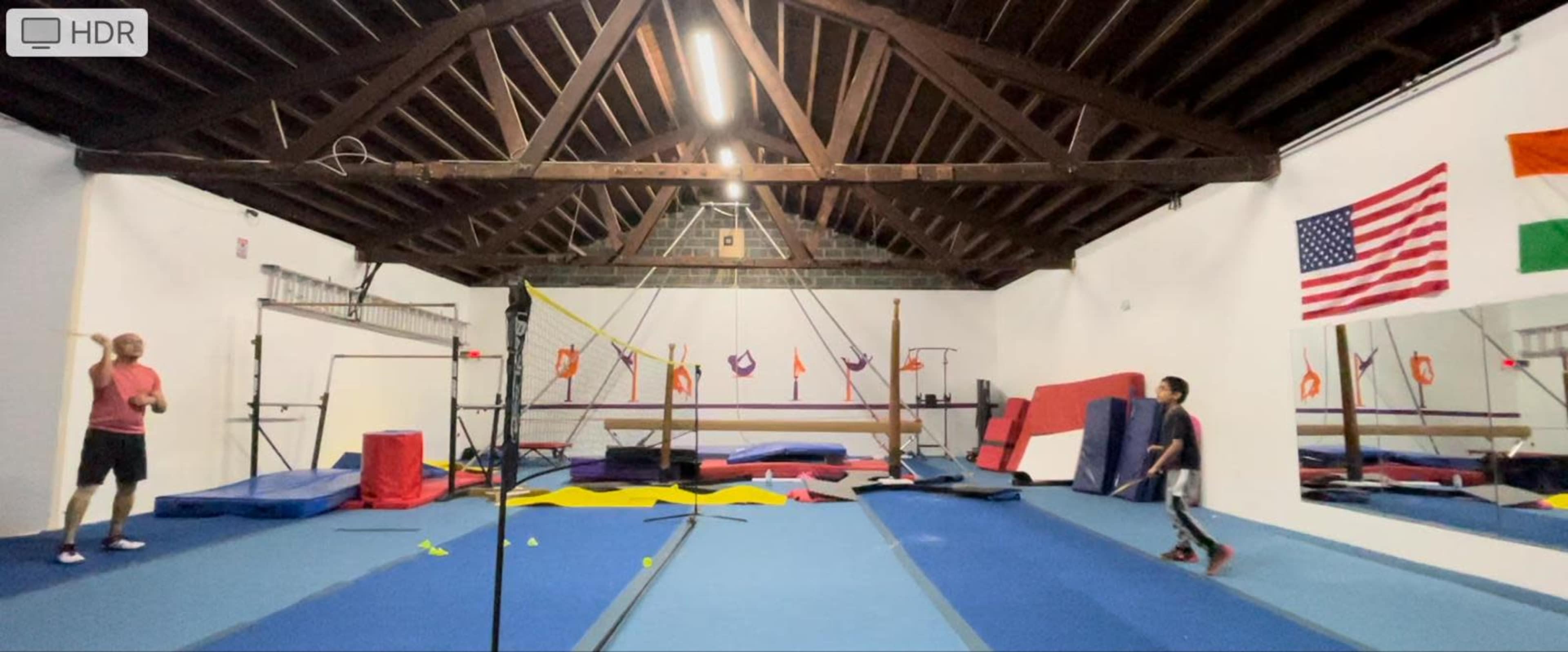 A spacious gymnasium equipped with gymnastics apparatus and colorful mats, with a person practicing on the left and another individual in motion on the right.