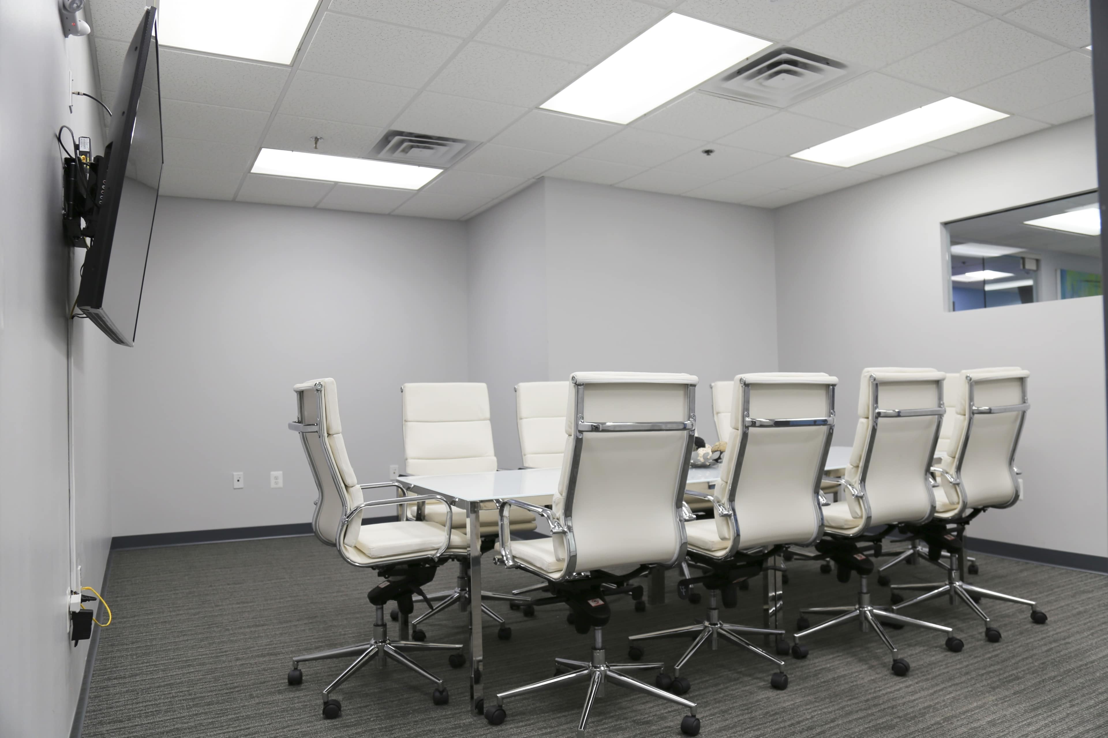 A modern conference room featuring a rectangular white table surrounded by eight white chairs and a wall-mounted television.