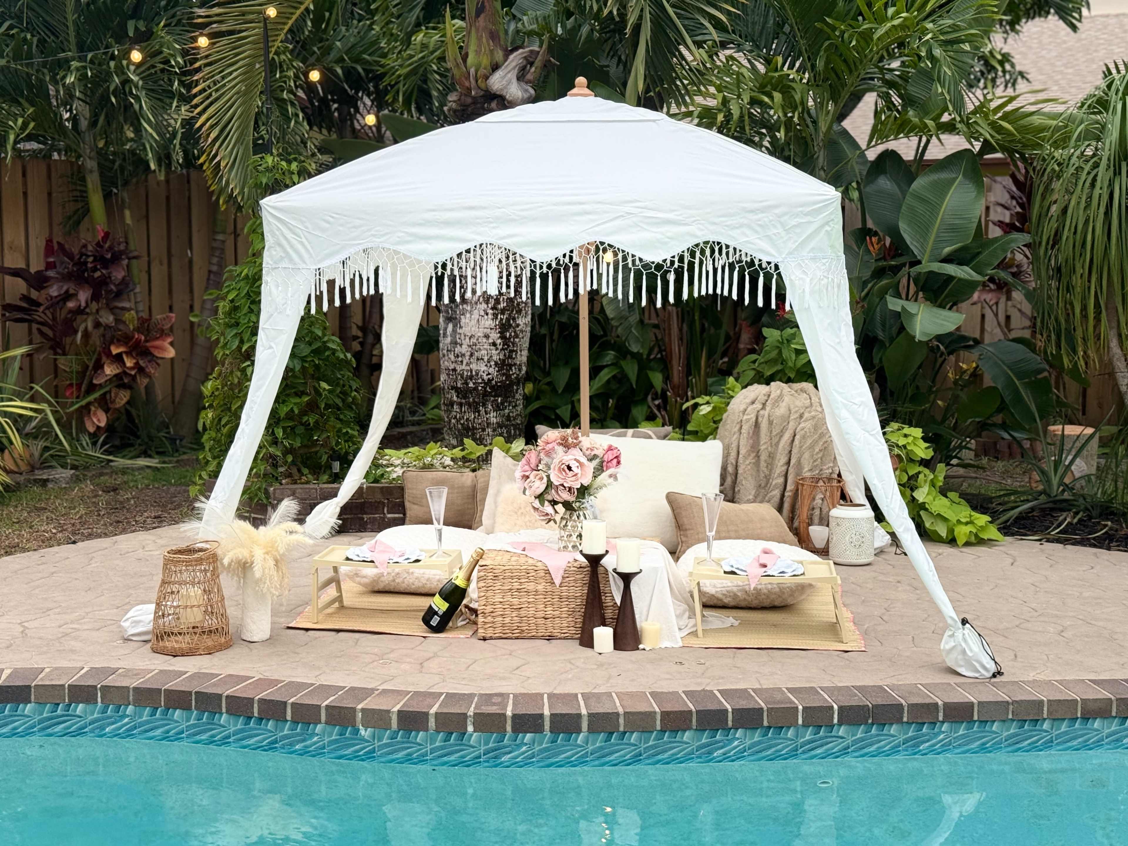 A white canopy covers a cozy outdoor seating area with decorative pillows, a floral arrangement, and a bottle of champagne near a swimming pool.