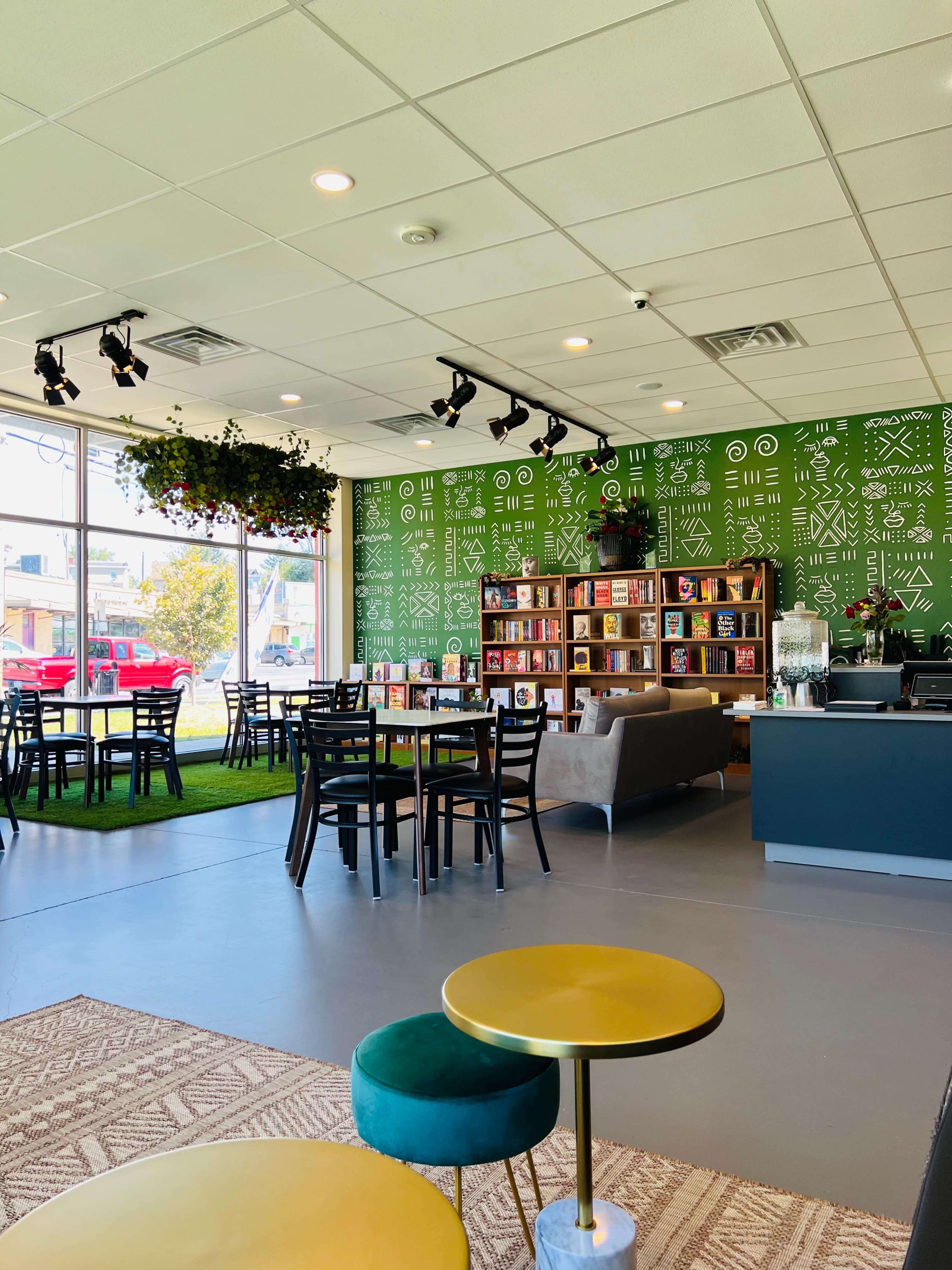 A modern cafe interior with green patterned walls, wooden bookshelves filled with books, tables and chairs, and a cozy seating area.