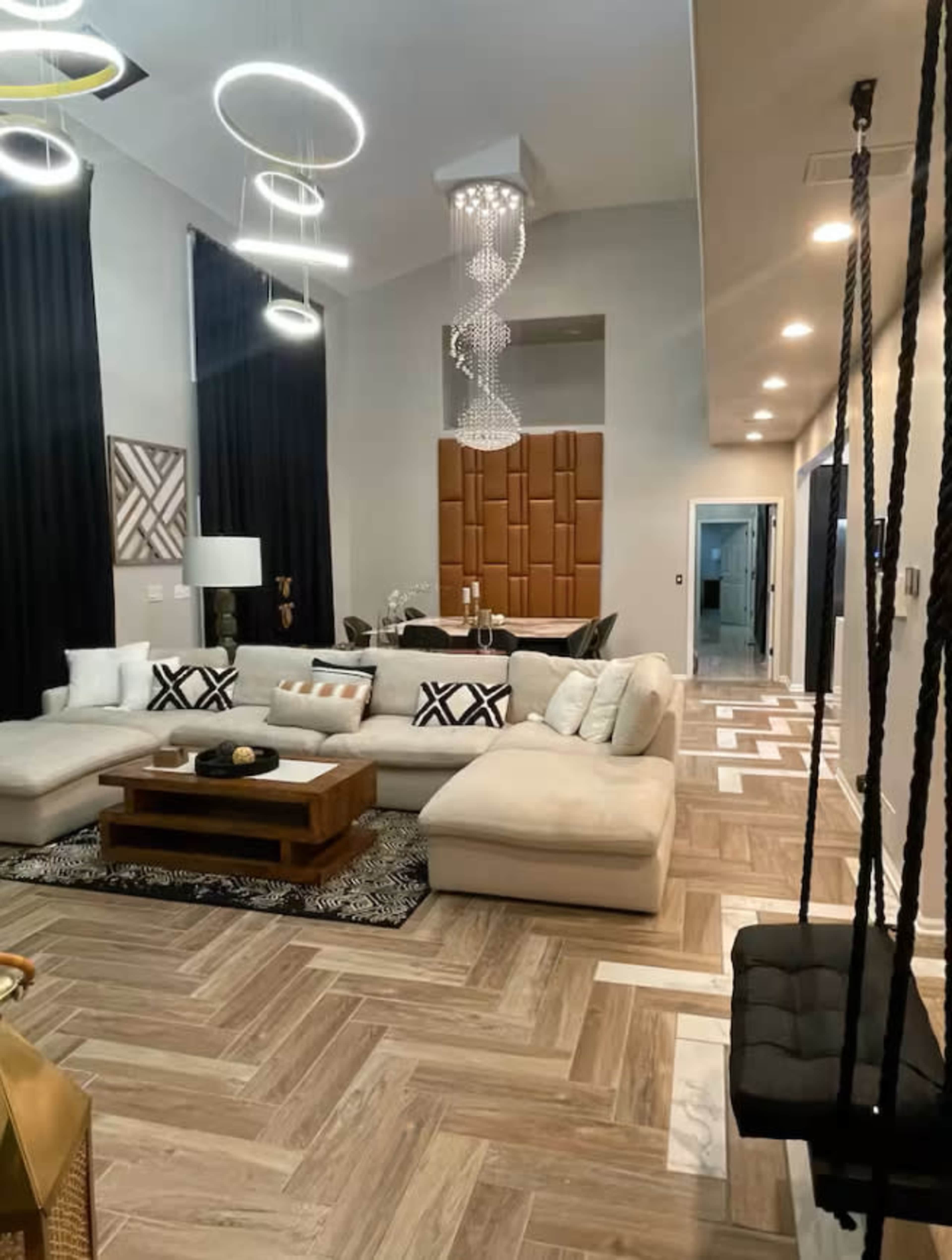 The image shows a stylish living room featuring a large sectional sofa, an elegant coffee table, modern lighting fixtures, and a dining area in the background.