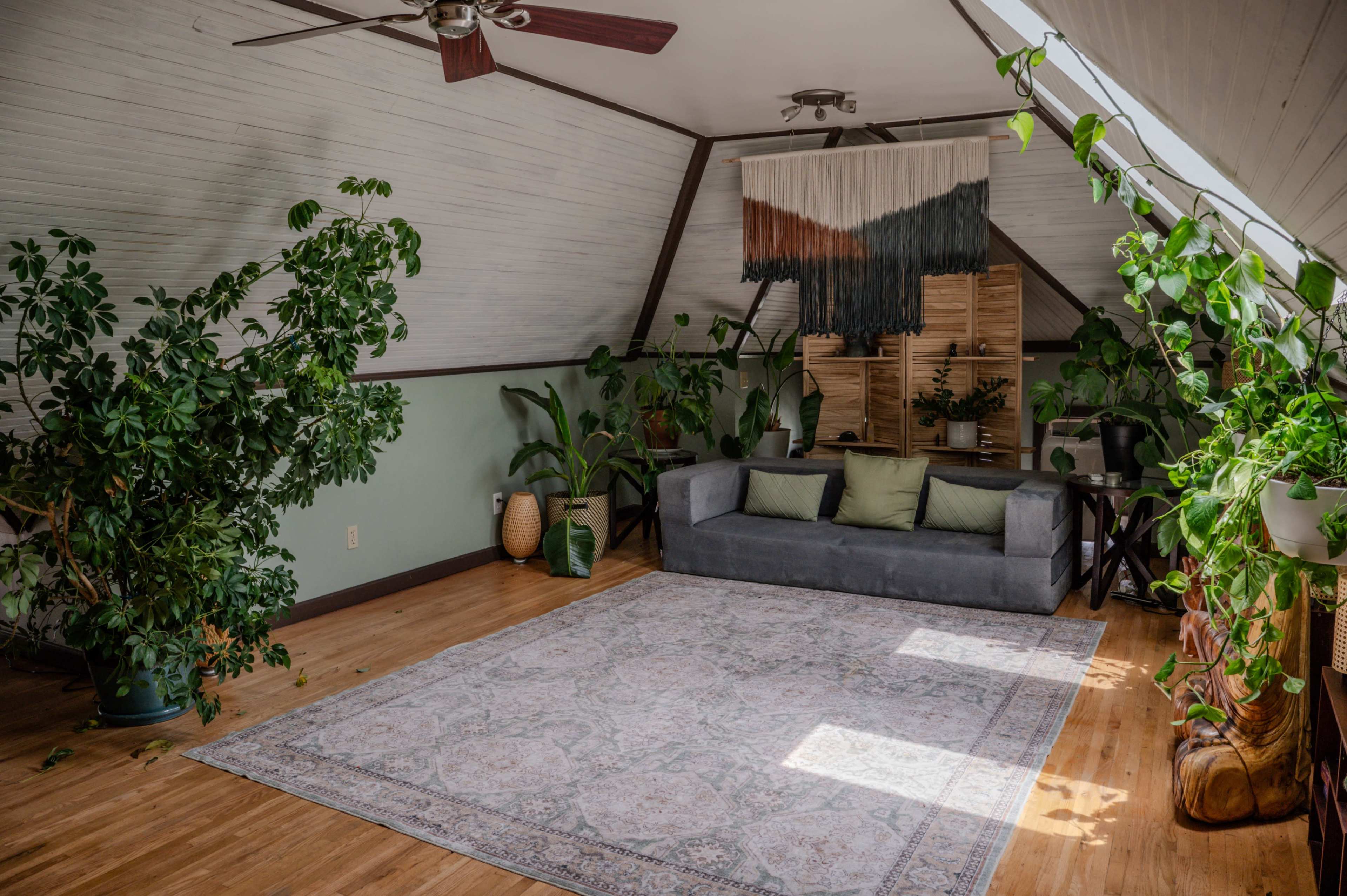 The image shows an attic room with wooden floors, a gray couch adorned with green pillows, plants throughout the space, and a decorative wall hanging.