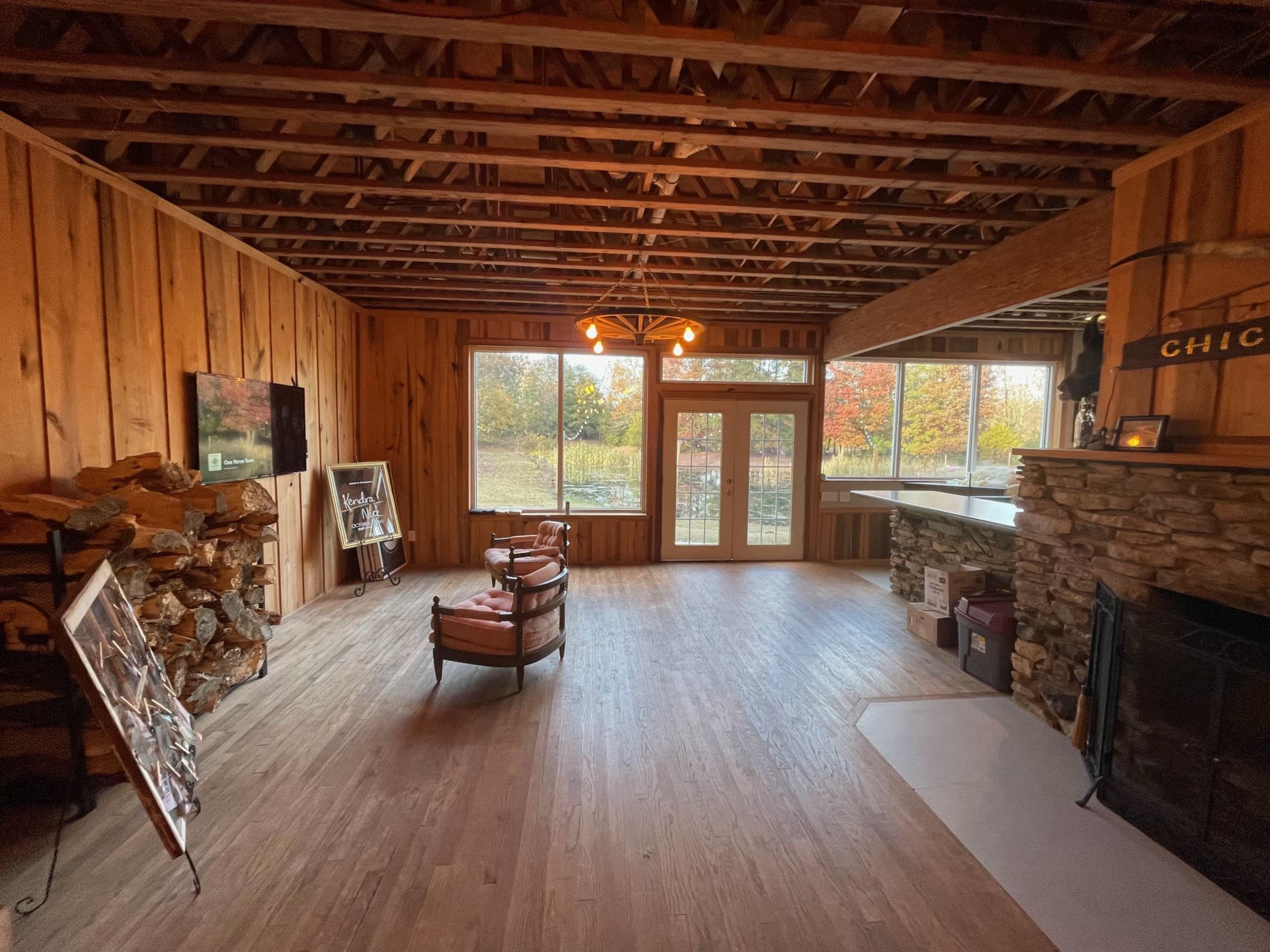A rustic interior with wooden walls, a stone fireplace, and large windows providing a view of an autumn landscape.