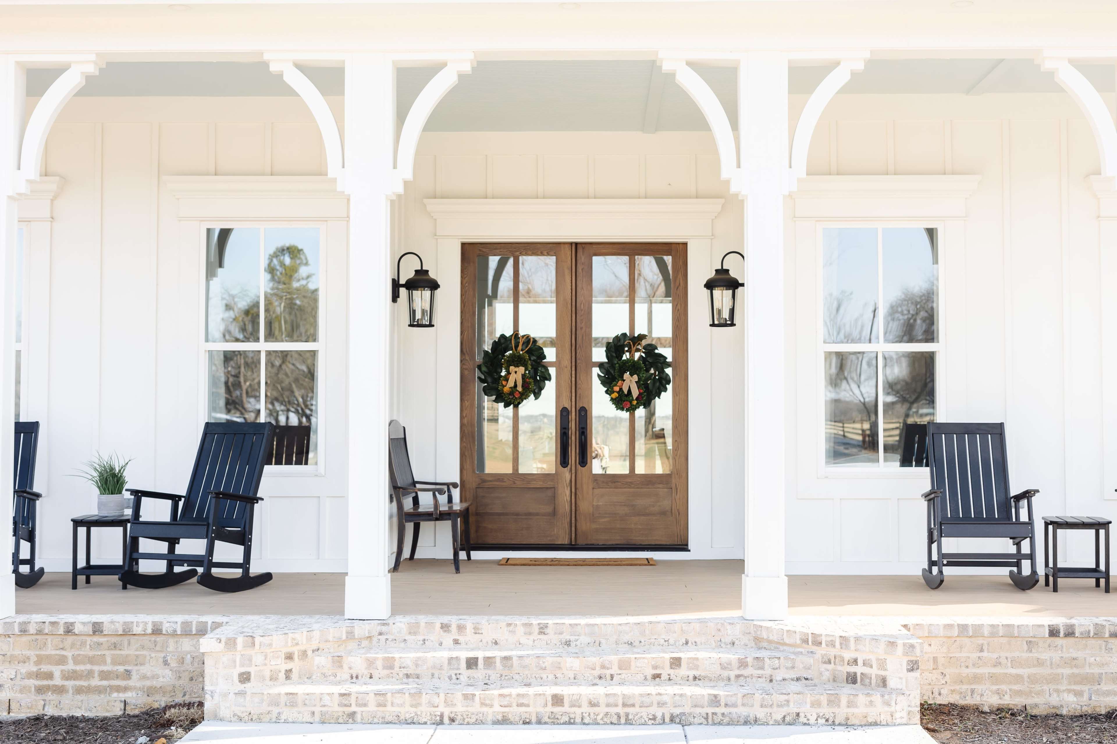The entrance features double wooden doors framed by two wreaths, surrounded by rocking chairs on a porch with brick steps.