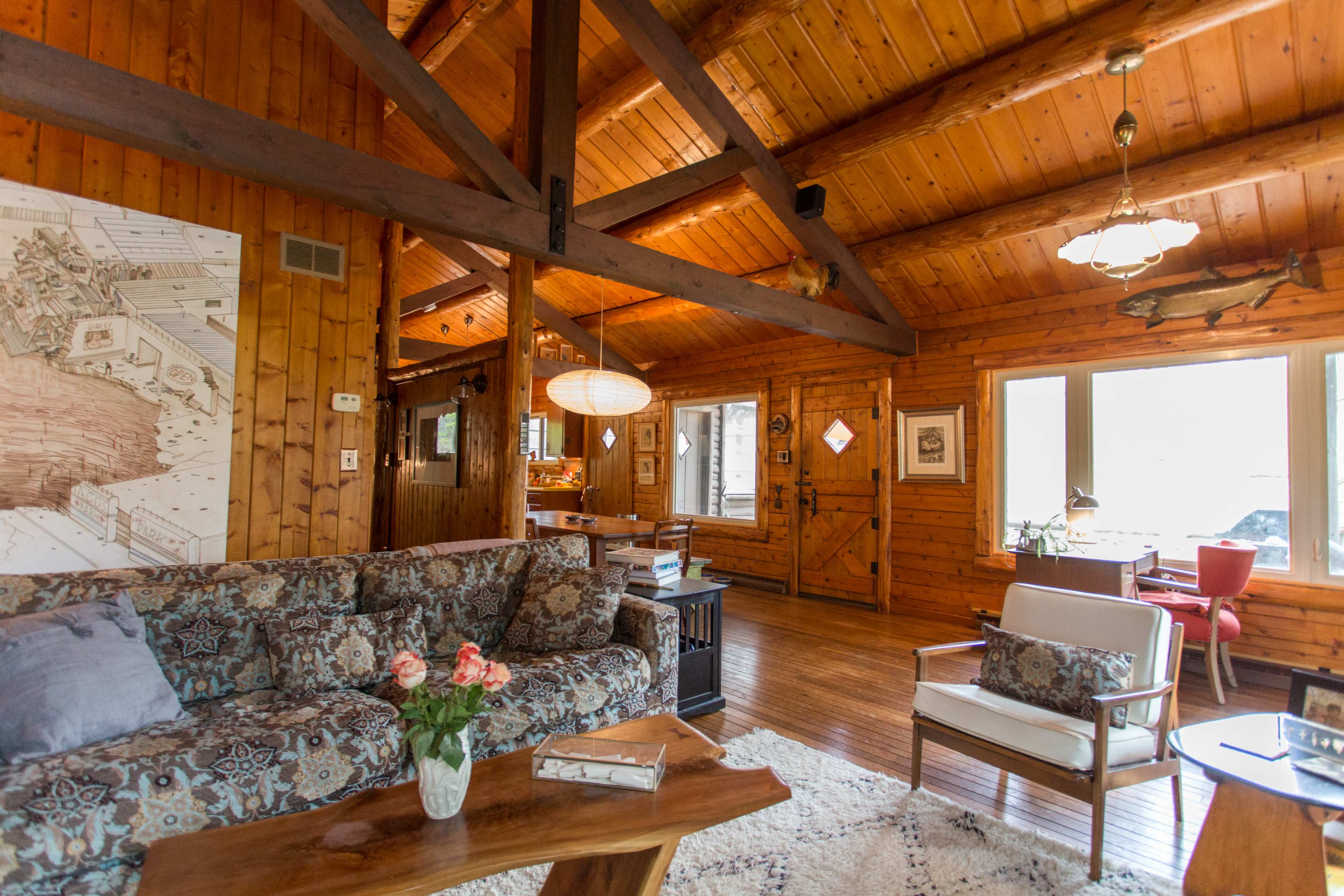 A wooden cabin interior with a combination of a living area featuring a patterned sofa, a coffee table, and large windows allowing natural light.