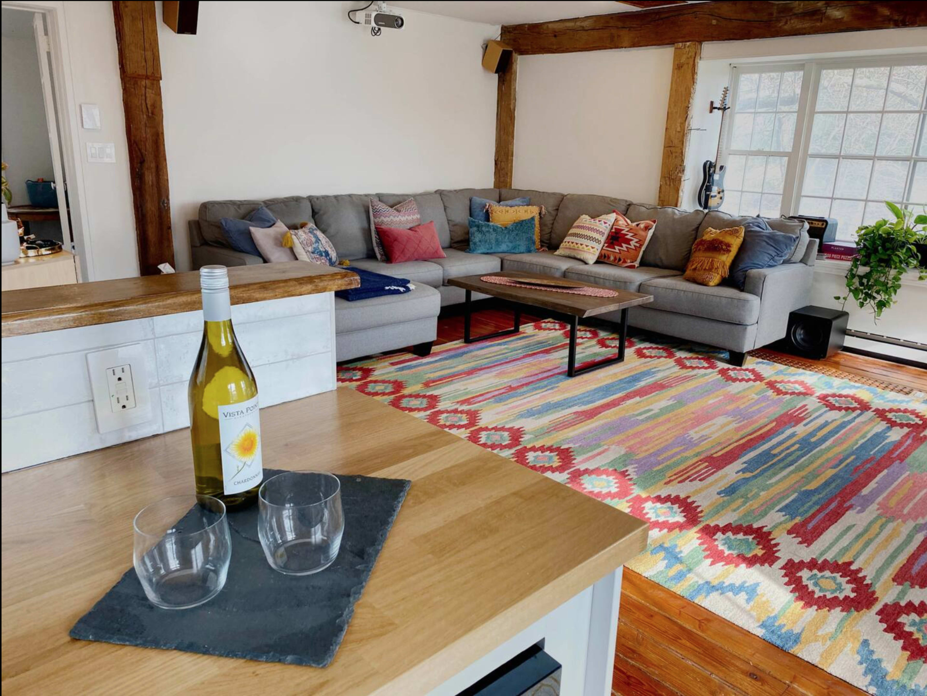 A cozy living room with a gray sectional sofa, colorful throw pillows, a wooden coffee table, and a patterned rug, along with a bottle of wine and two glasses on a wooden countertop.