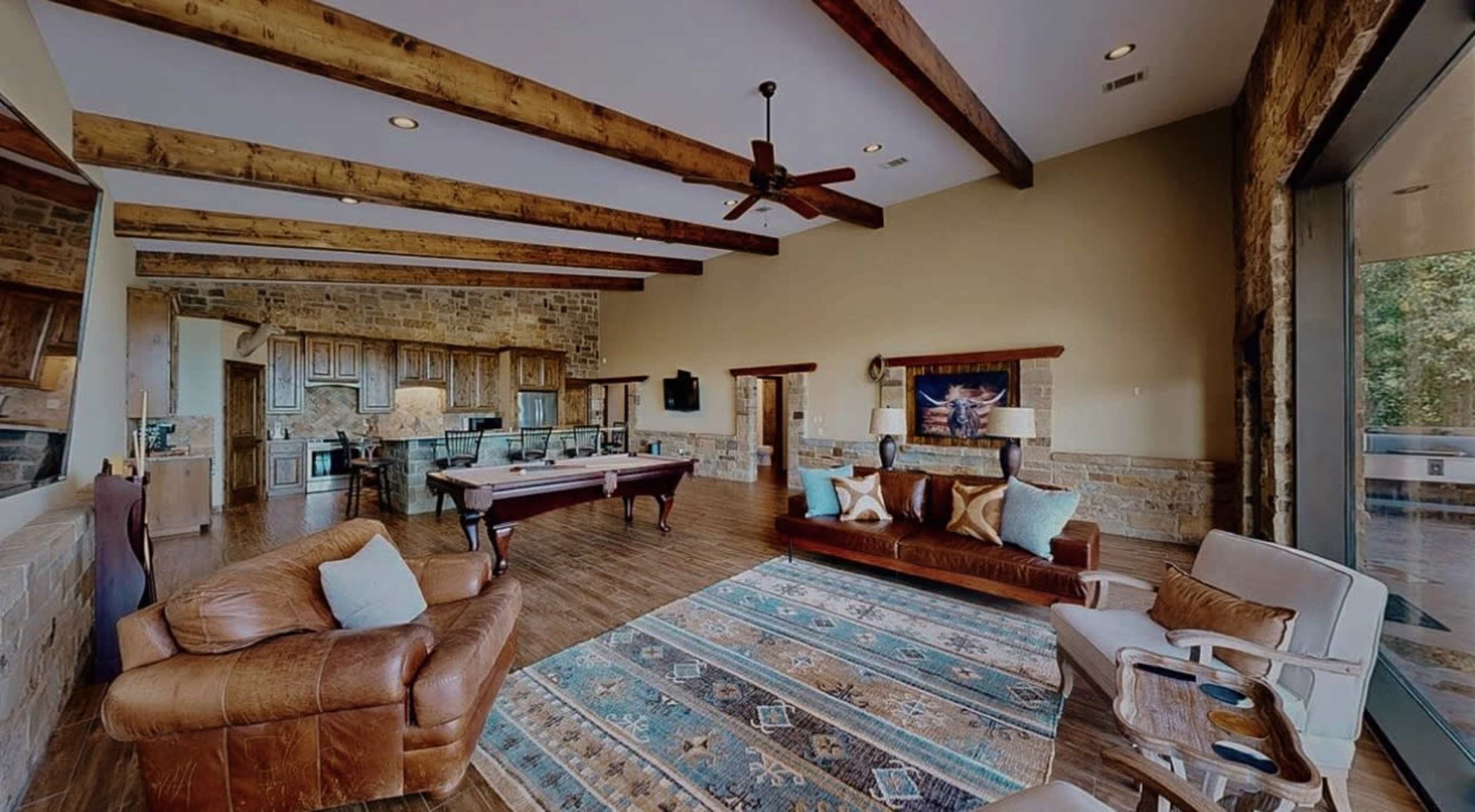 The image shows a spacious living area with wooden beams, a pool table, and a mix of seating options, including a leather sofa and armchairs, set against stone walls.