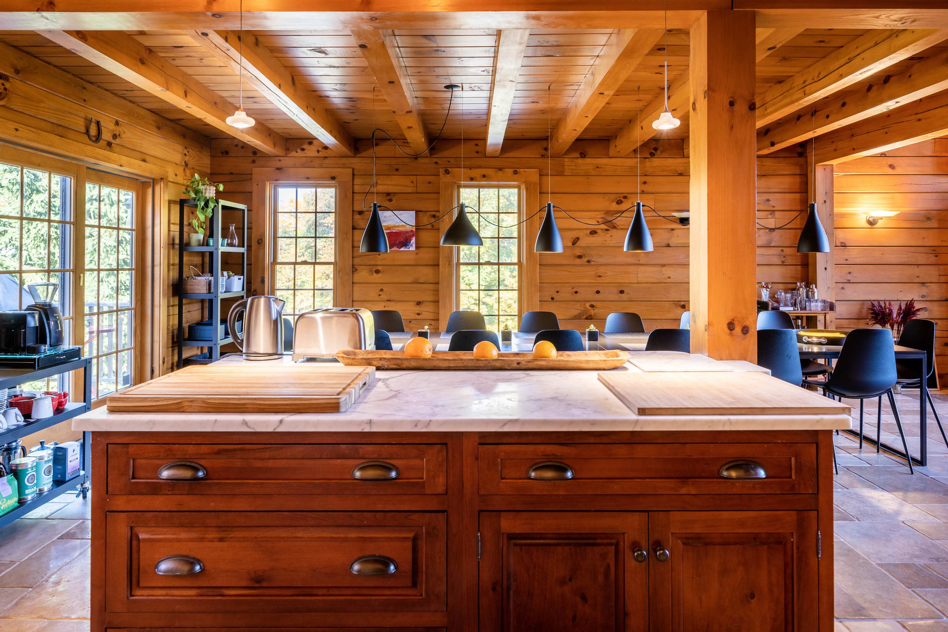 A spacious wooden kitchen features a marble-topped island, cabinets below, and a dining area with a large window.