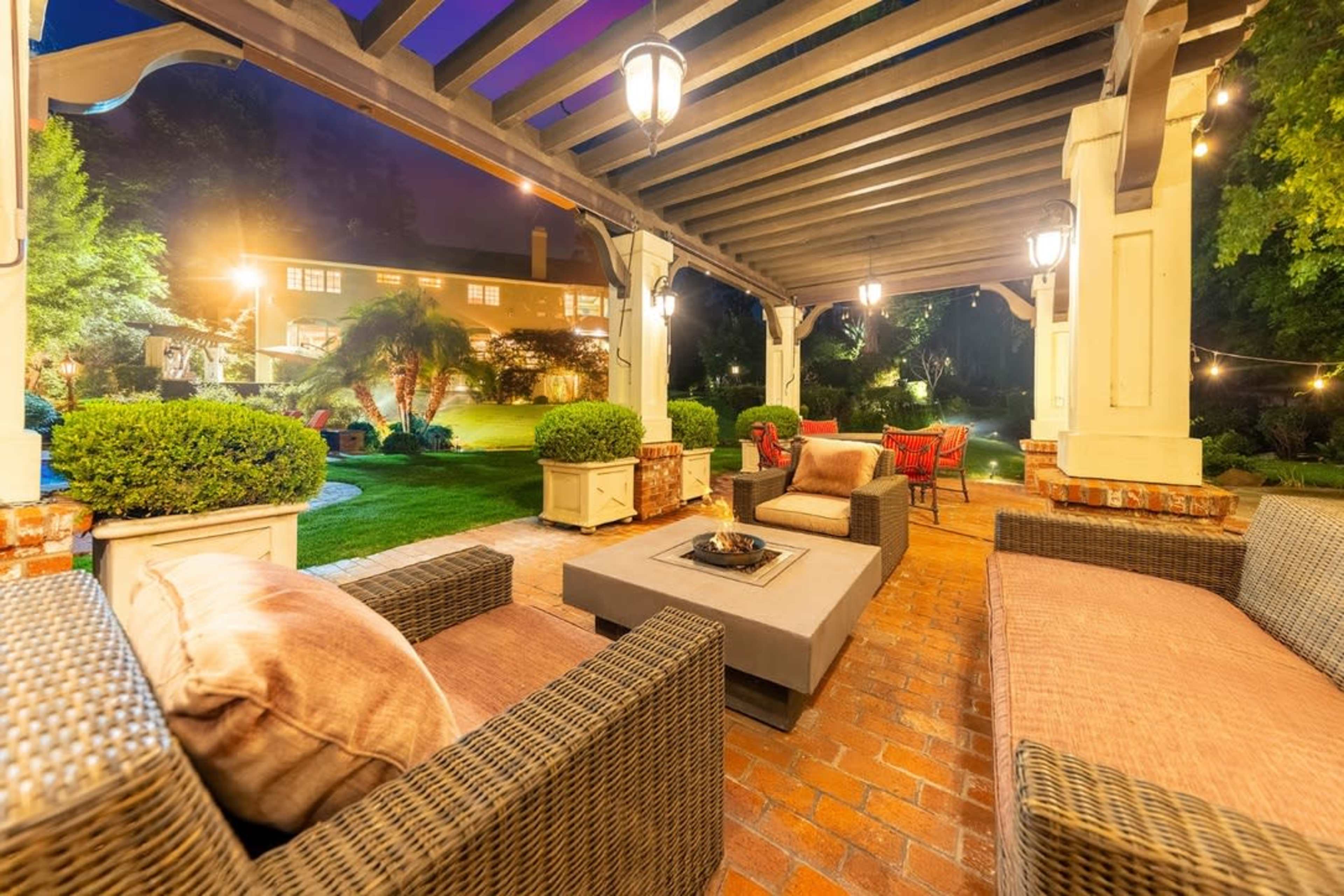 A covered patio area features wicker furniture arranged around a central fire pit, with a well-lit garden and a house visible in the background.