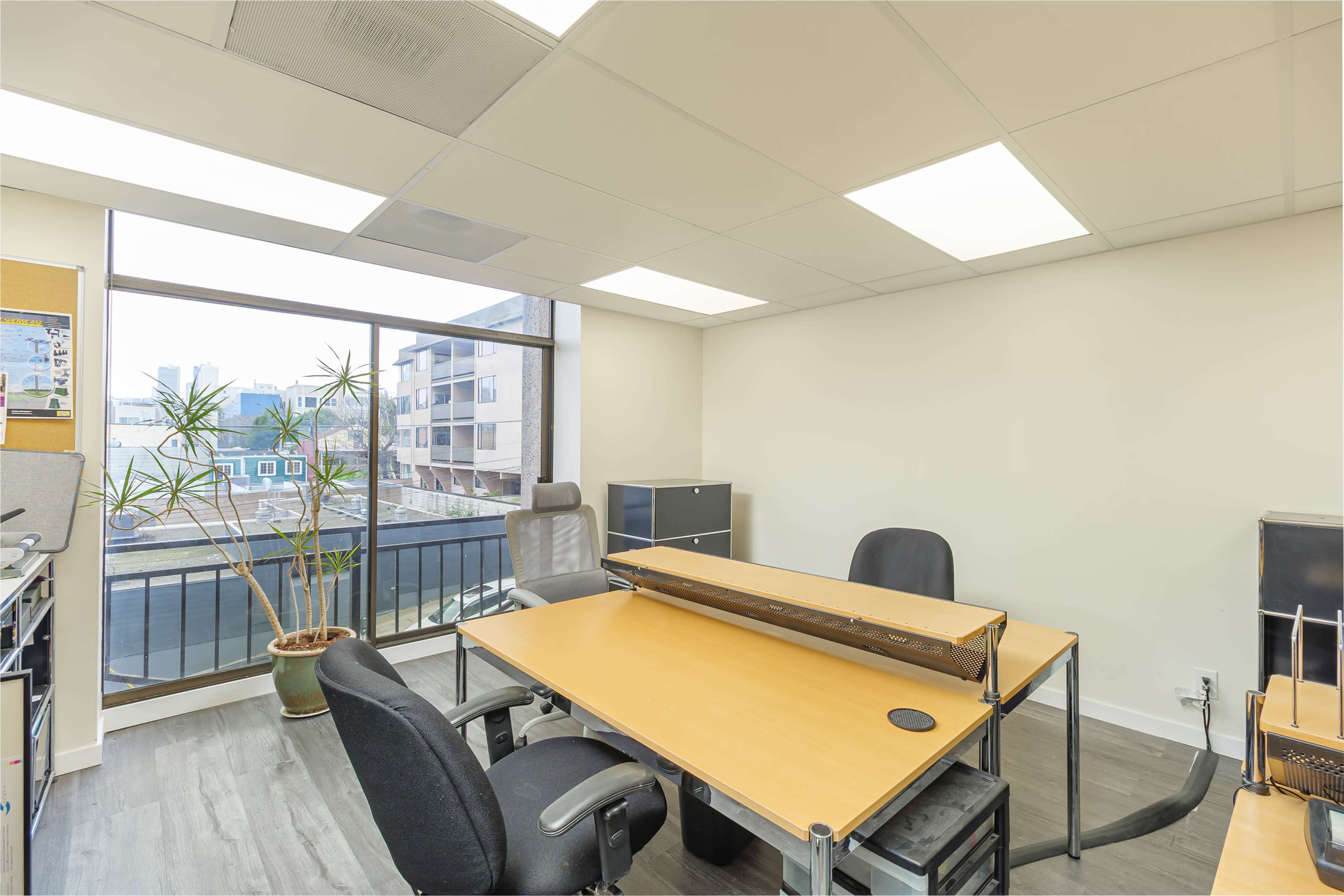 The image shows a modern office space with two desks and chairs, large windows overlooking a cityscape, and a potted plant in the corner.