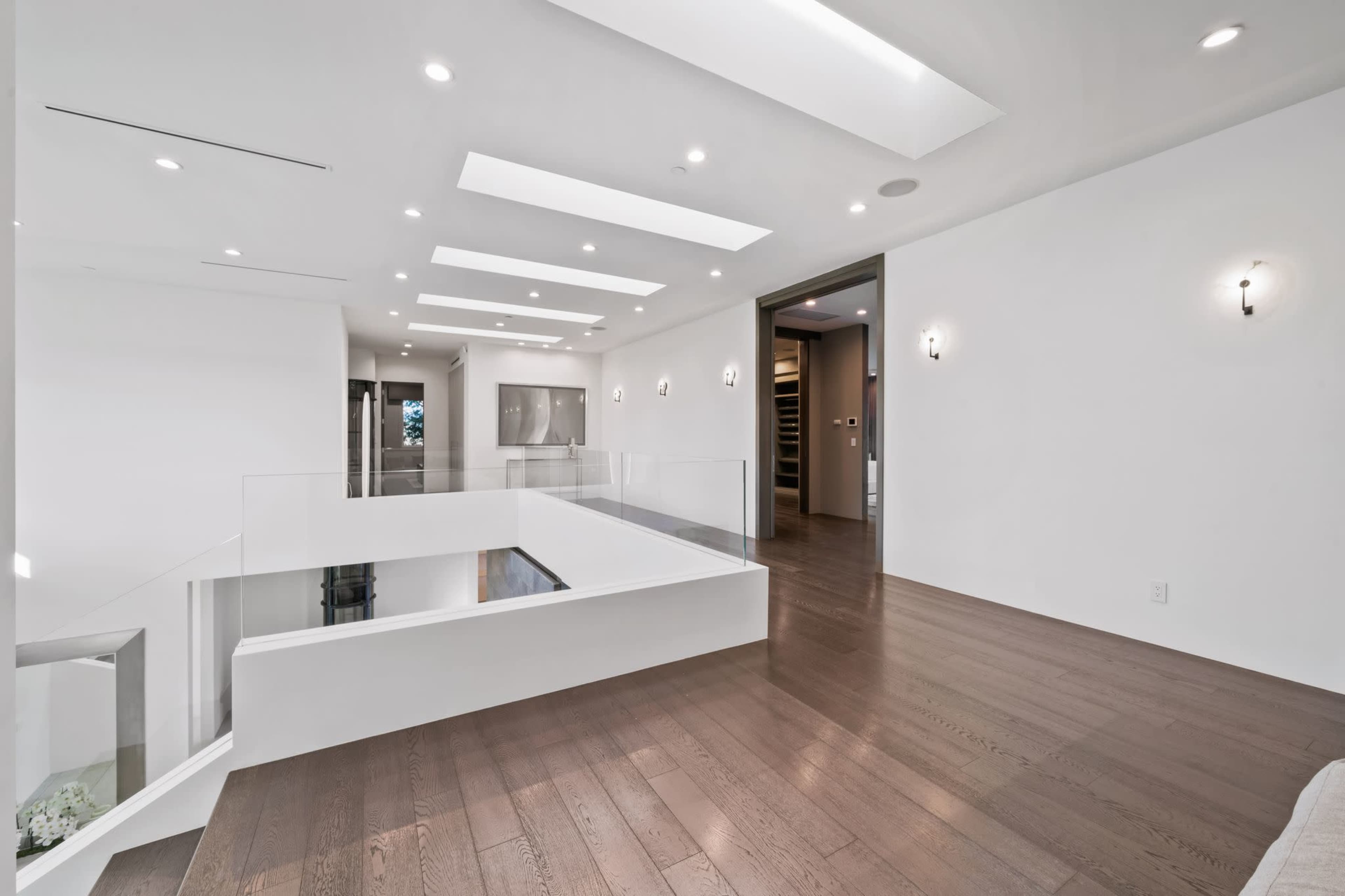 The image shows a modern interior of a house featuring a spacious, well-lit hallway with a wooden floor and a balcony railing overlooking a lower level.
