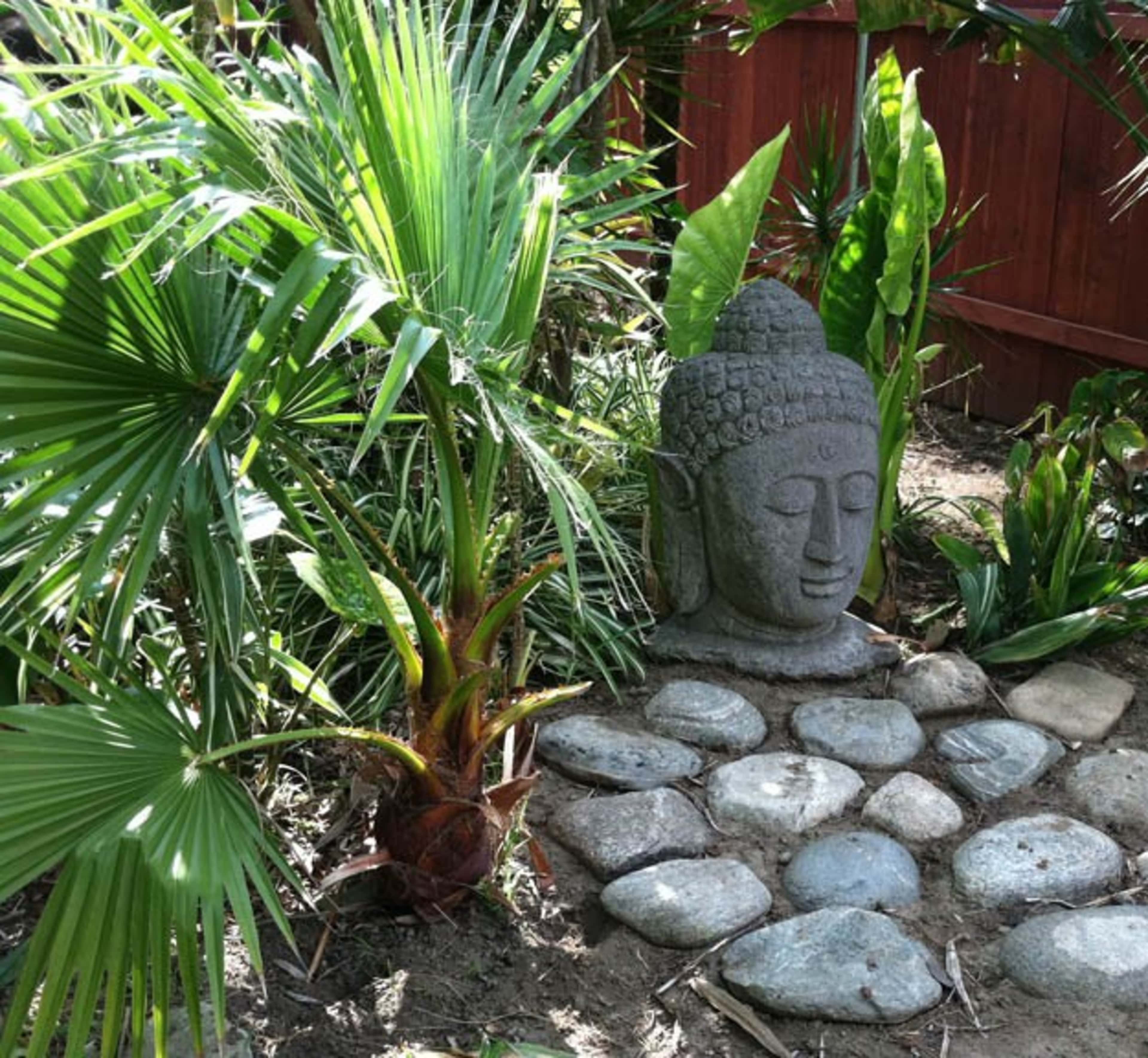 A stone Buddha statue is surrounded by greenery and smooth stones in a garden setting.
