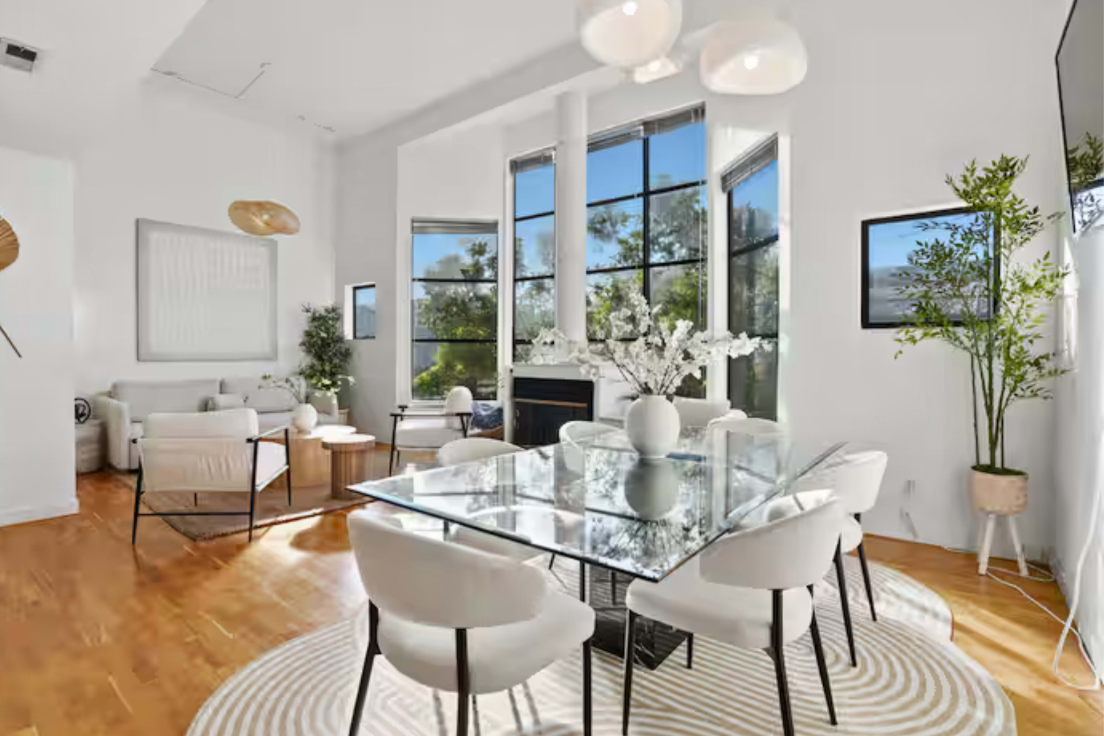 The image shows a bright, modern dining area with a glass table, white chairs, and large windows allowing natural light to fill the space, overlooking greenery.