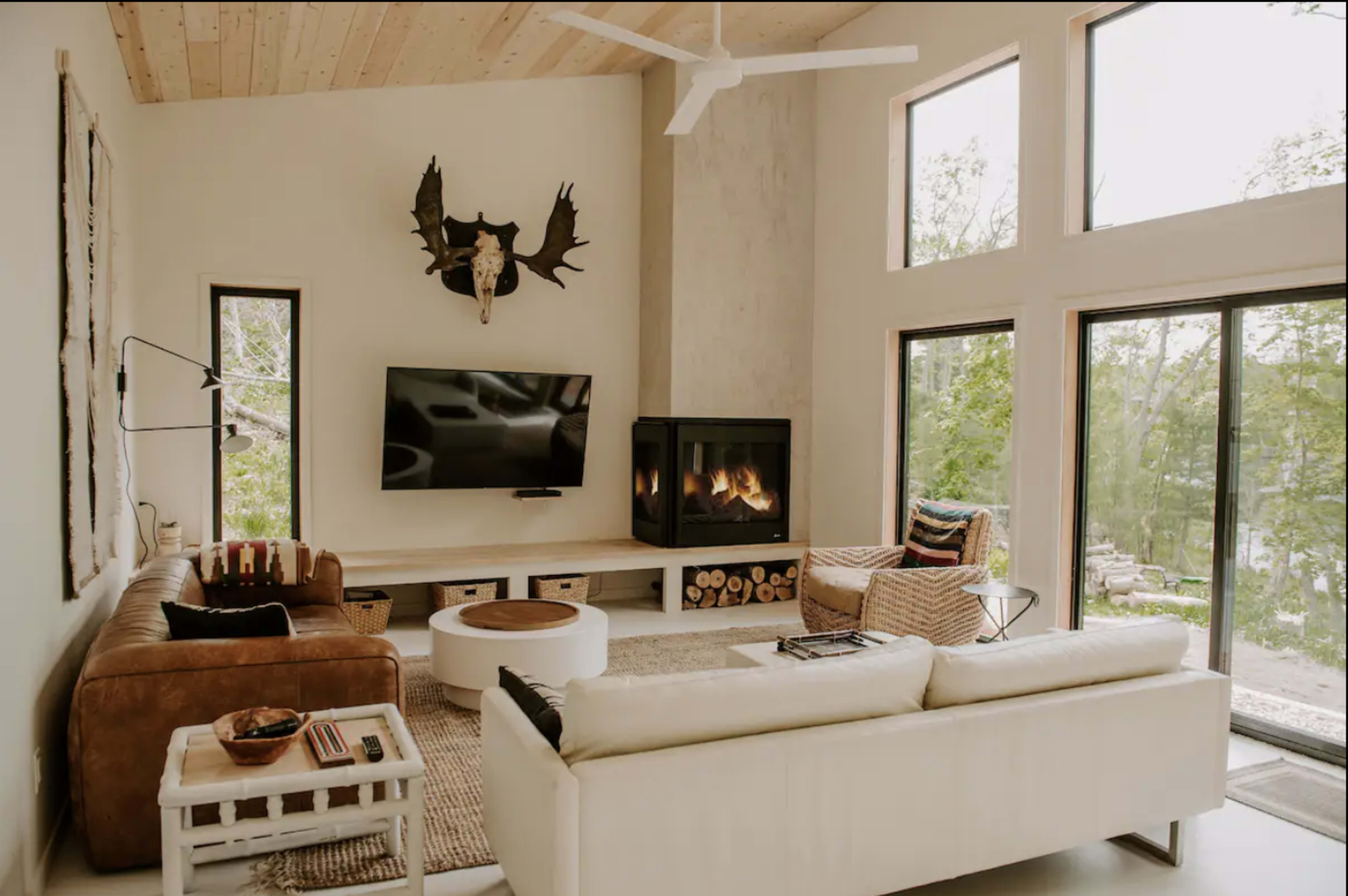 A modern living room features large windows, a mounted moose head, a flat-screen TV, a fireplace, and both leather and white sofas.