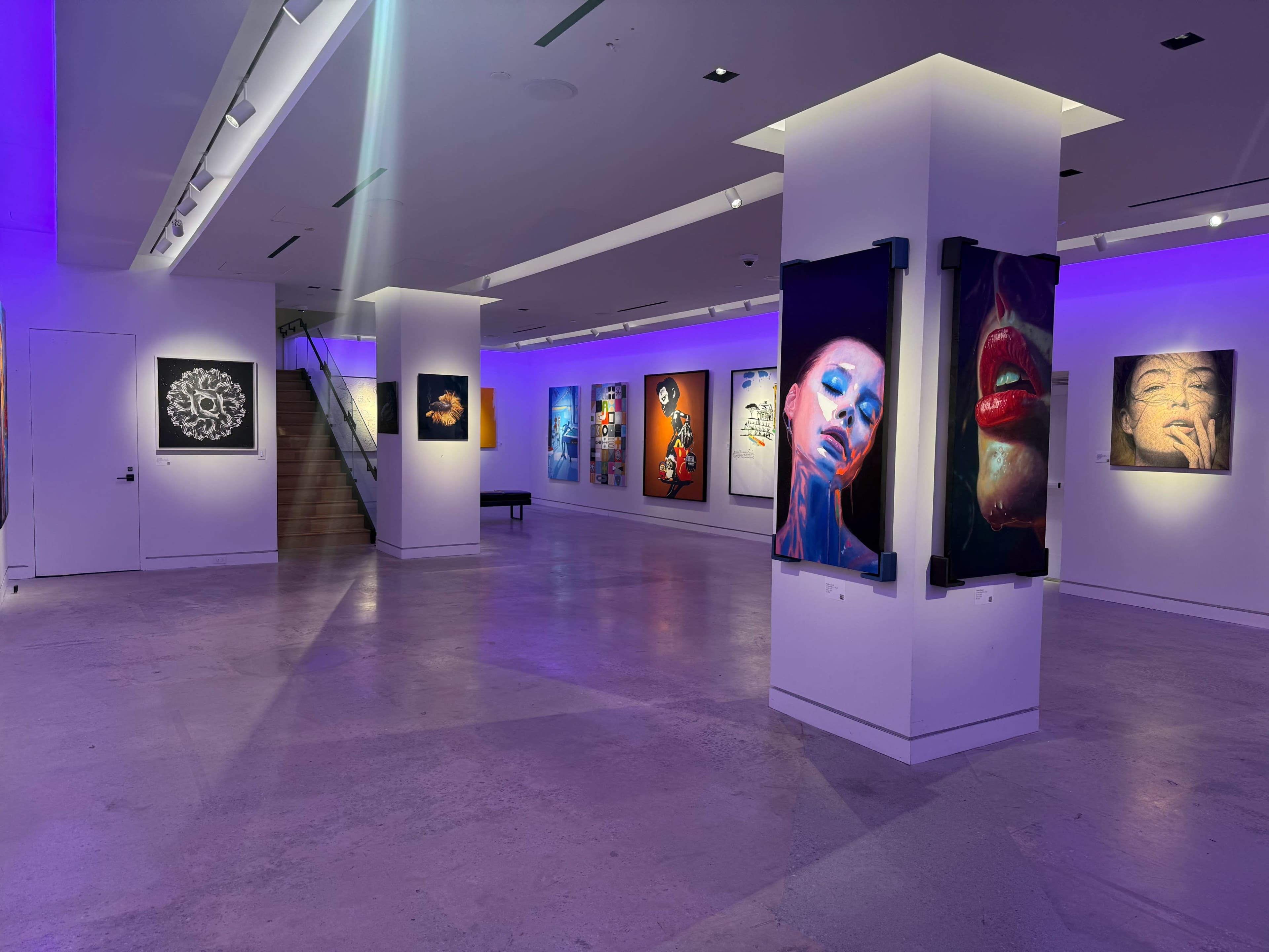 The image shows a modern art gallery featuring various colorful artworks displayed on the walls, illuminated by purple lighting.