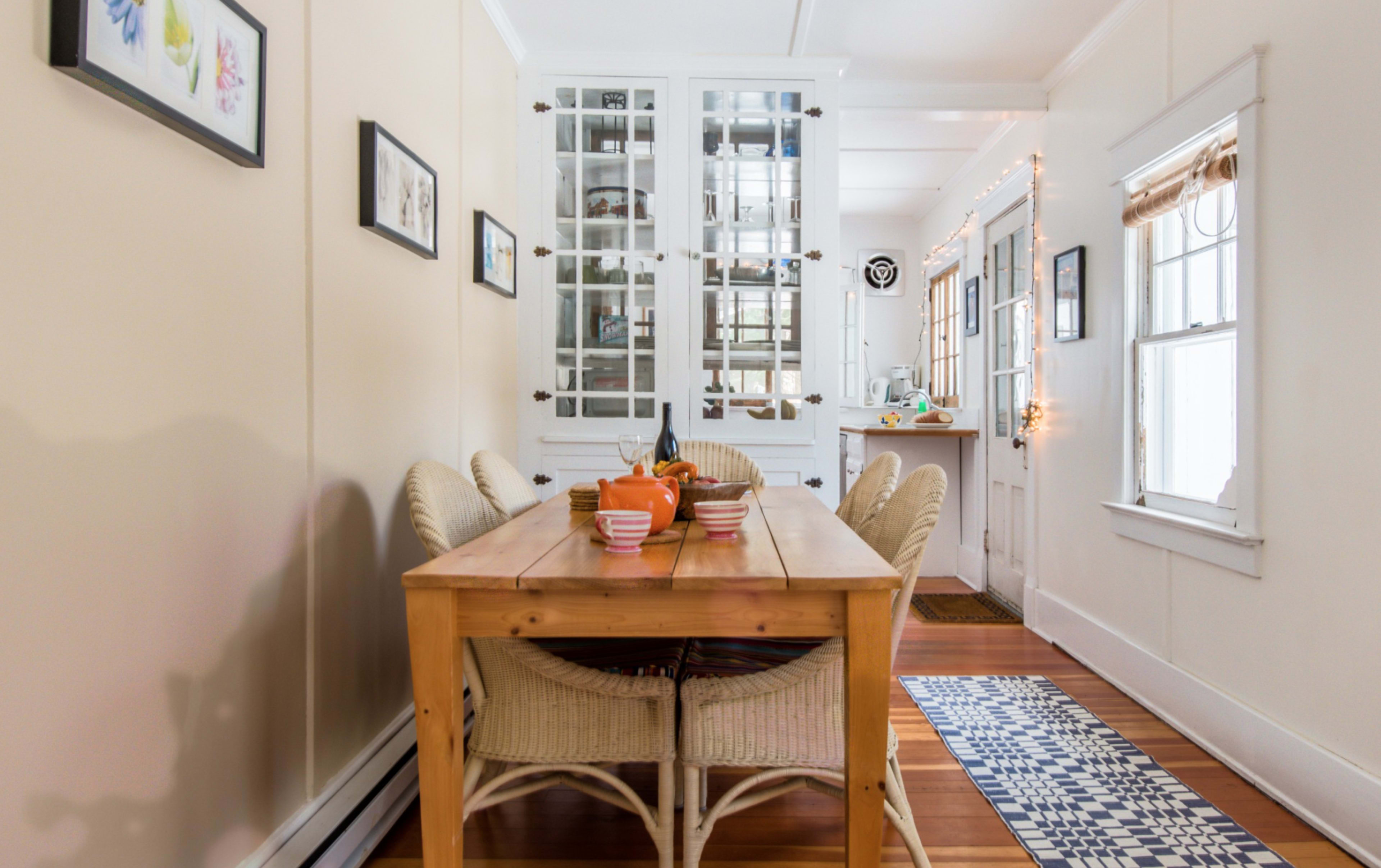 A wooden dining table with striped bowls filled with fruit is surrounded by wicker chairs in a bright, narrow room featuring white walls and large windows.
