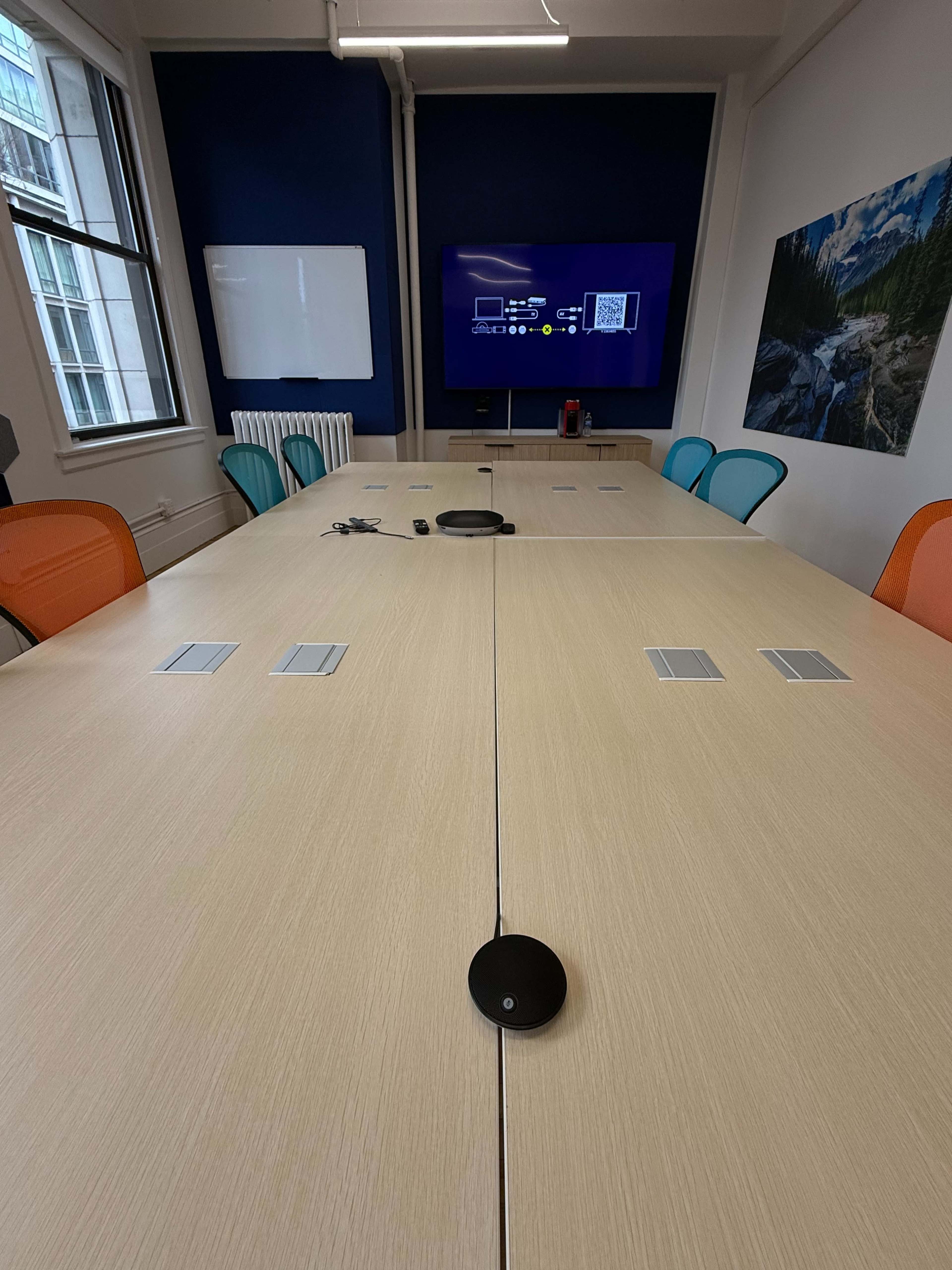 A conference room with a long wooden table, equipped with multiple charging ports, surrounded by colorful chairs, and a wall-mounted screen displaying a digital interface.