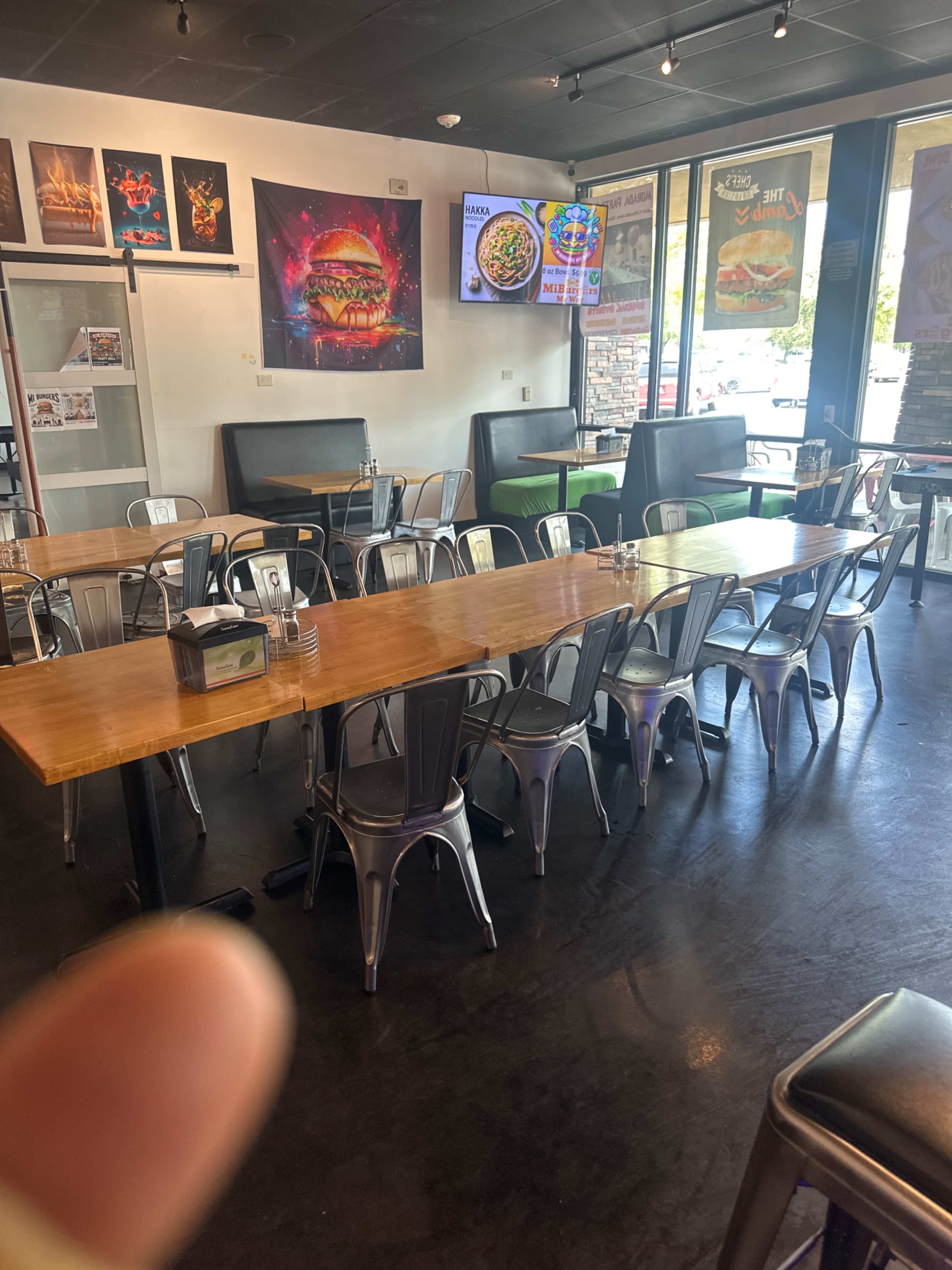 The image shows an interior view of a restaurant with metal and wooden tables, benches, and large windows displaying colorful food posters.