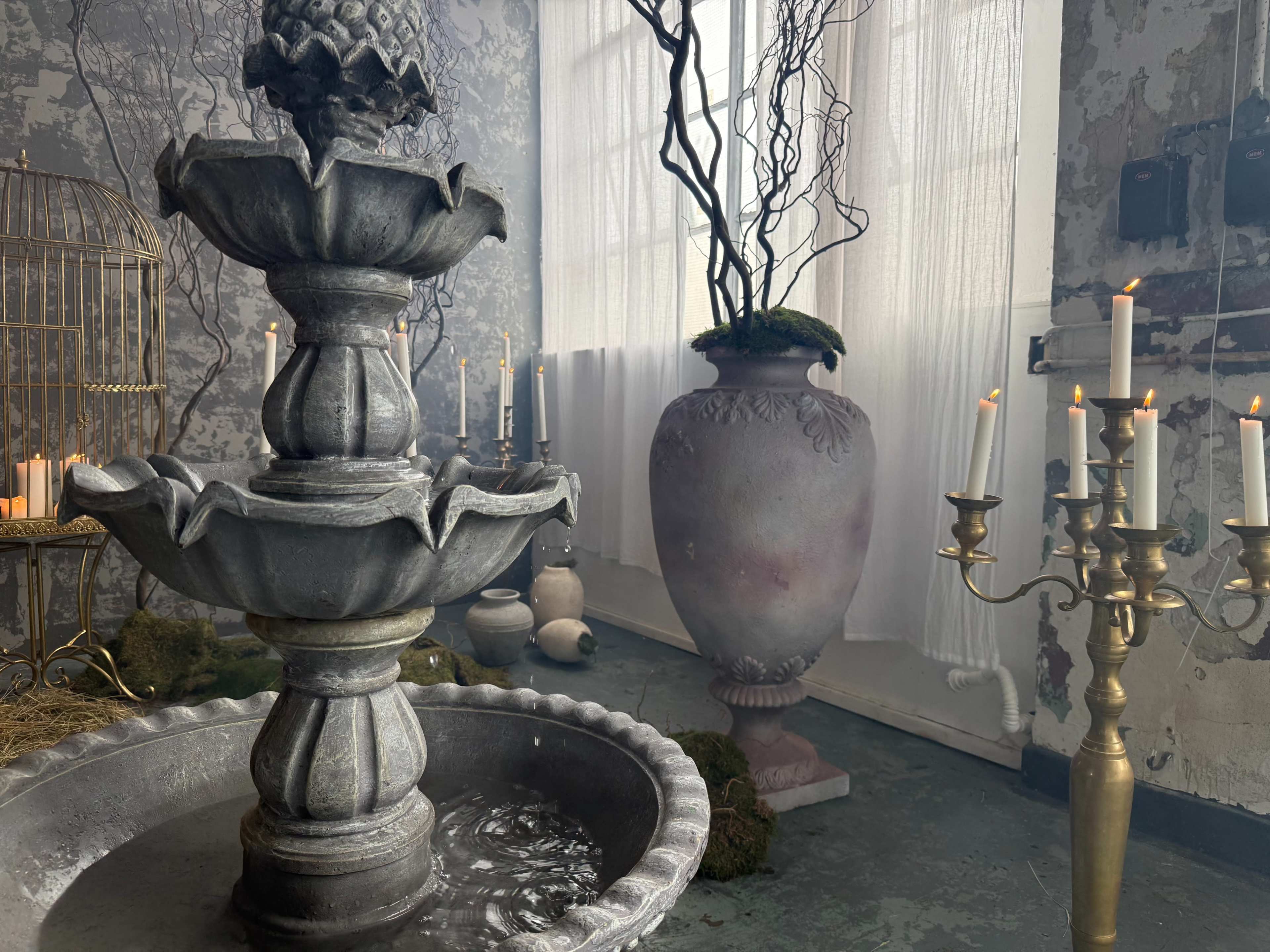 The image features a decorative stone fountain in an indoor space adorned with an ornate vase, a birdcage, and candles on a candelabrum.