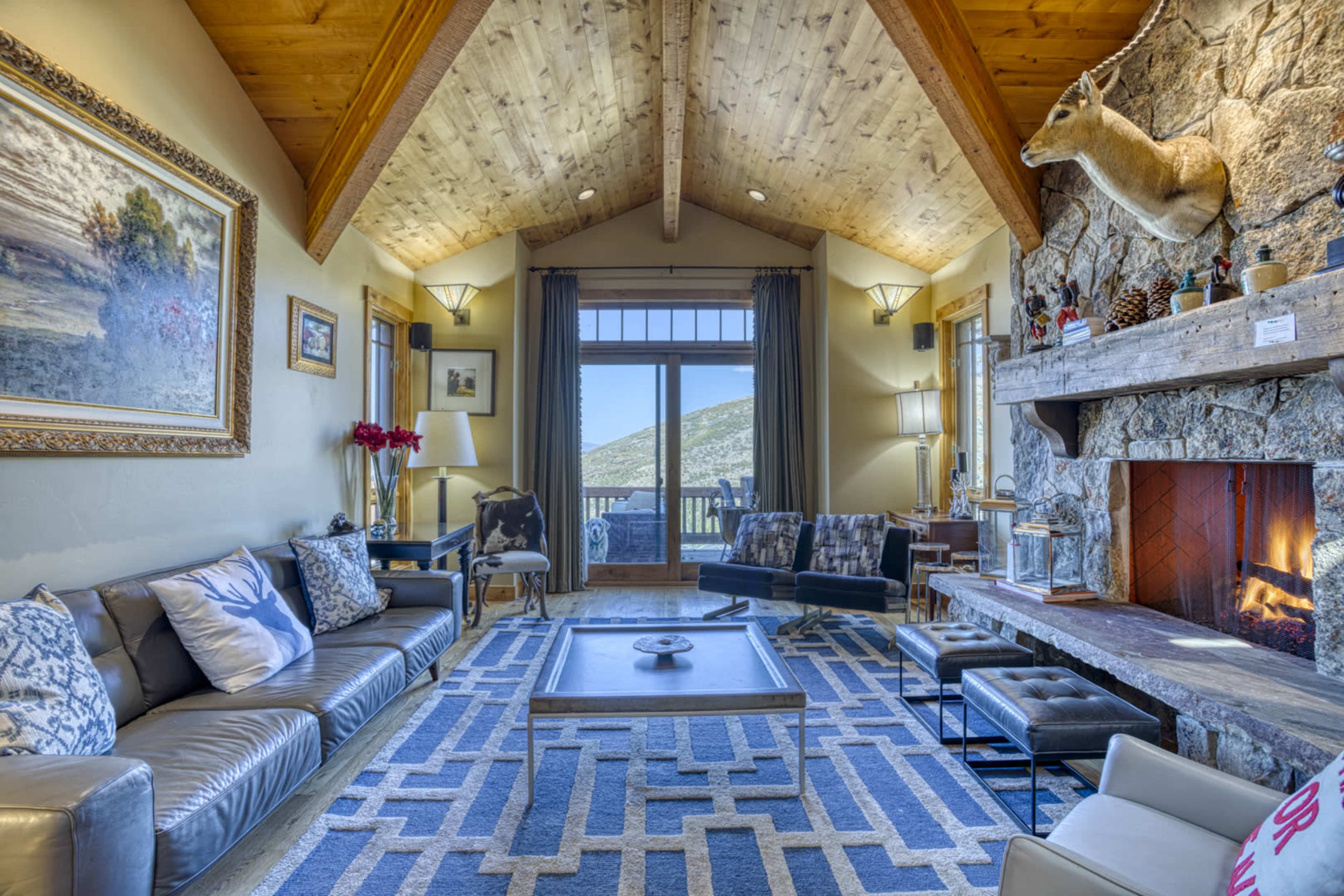 A cozy living room with a stone fireplace, wooden beams, and large windows offering a scenic view.