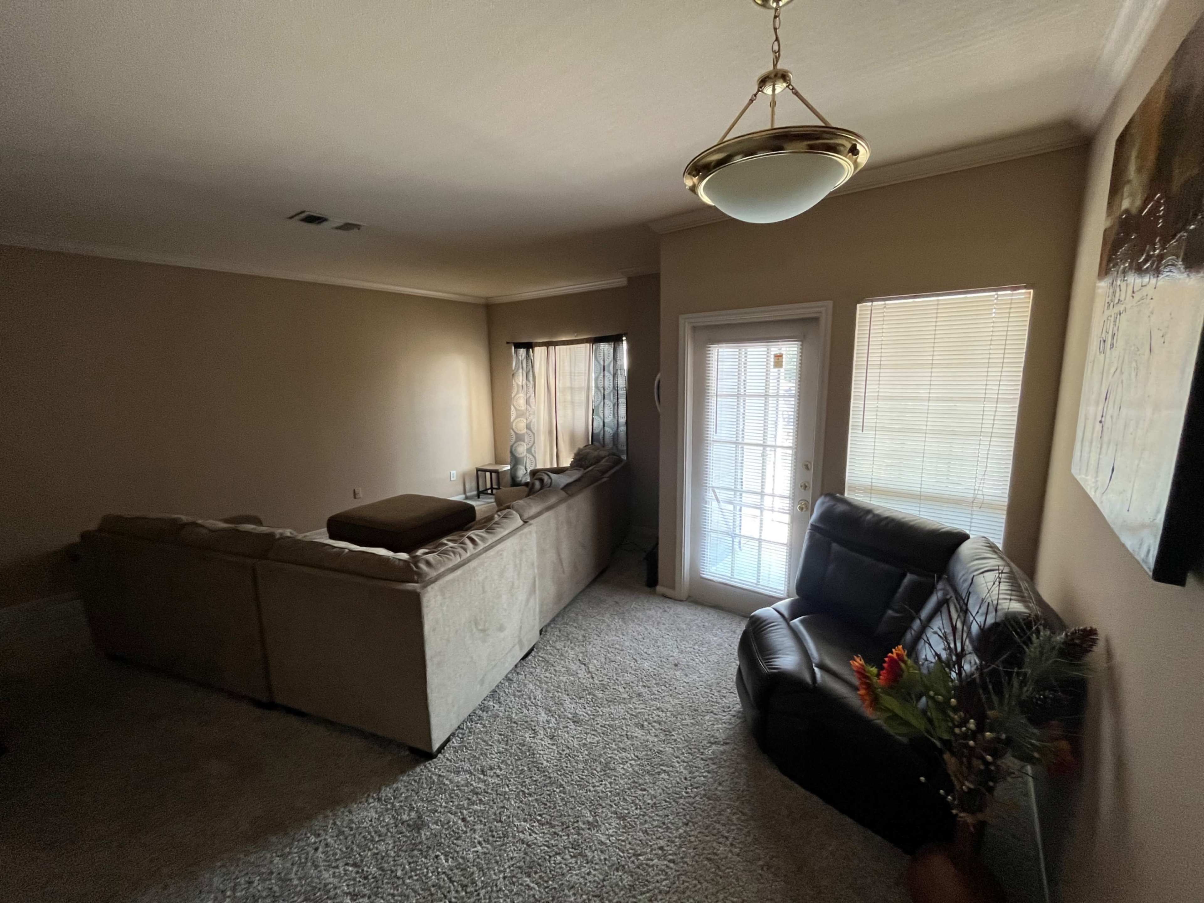 A spacious living room features a beige sectional sofa, a dark leather chair, and sliding glass doors leading to an outdoor area.
