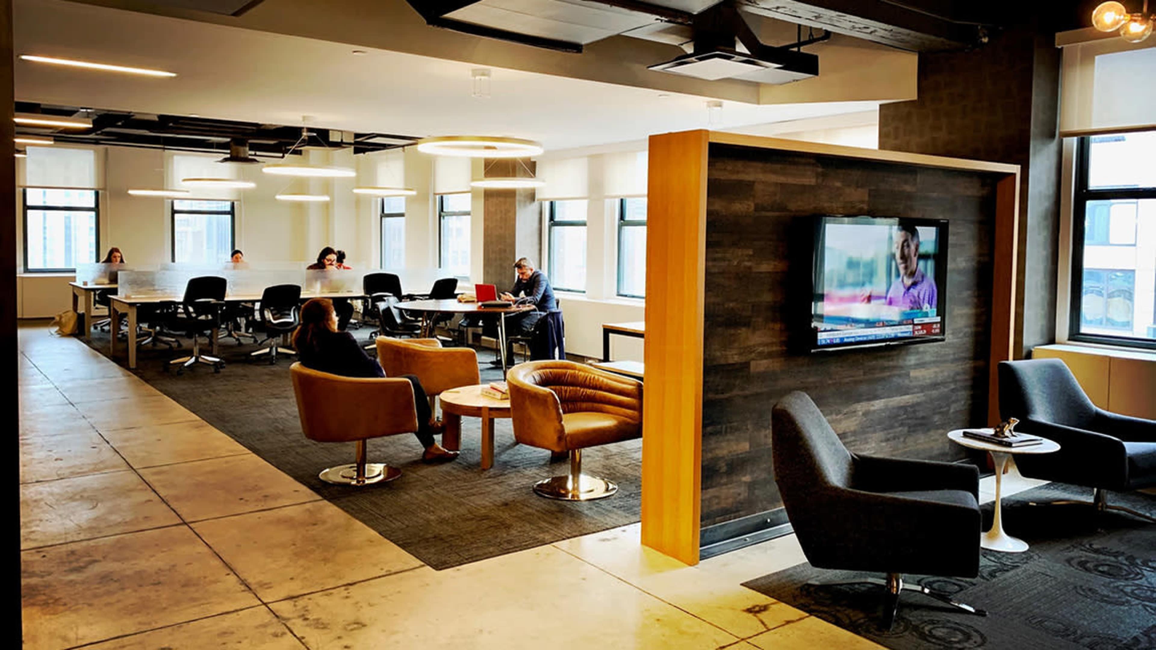 An open office space features various seating arrangements and a large table, with a television mounted on a wooden partition.