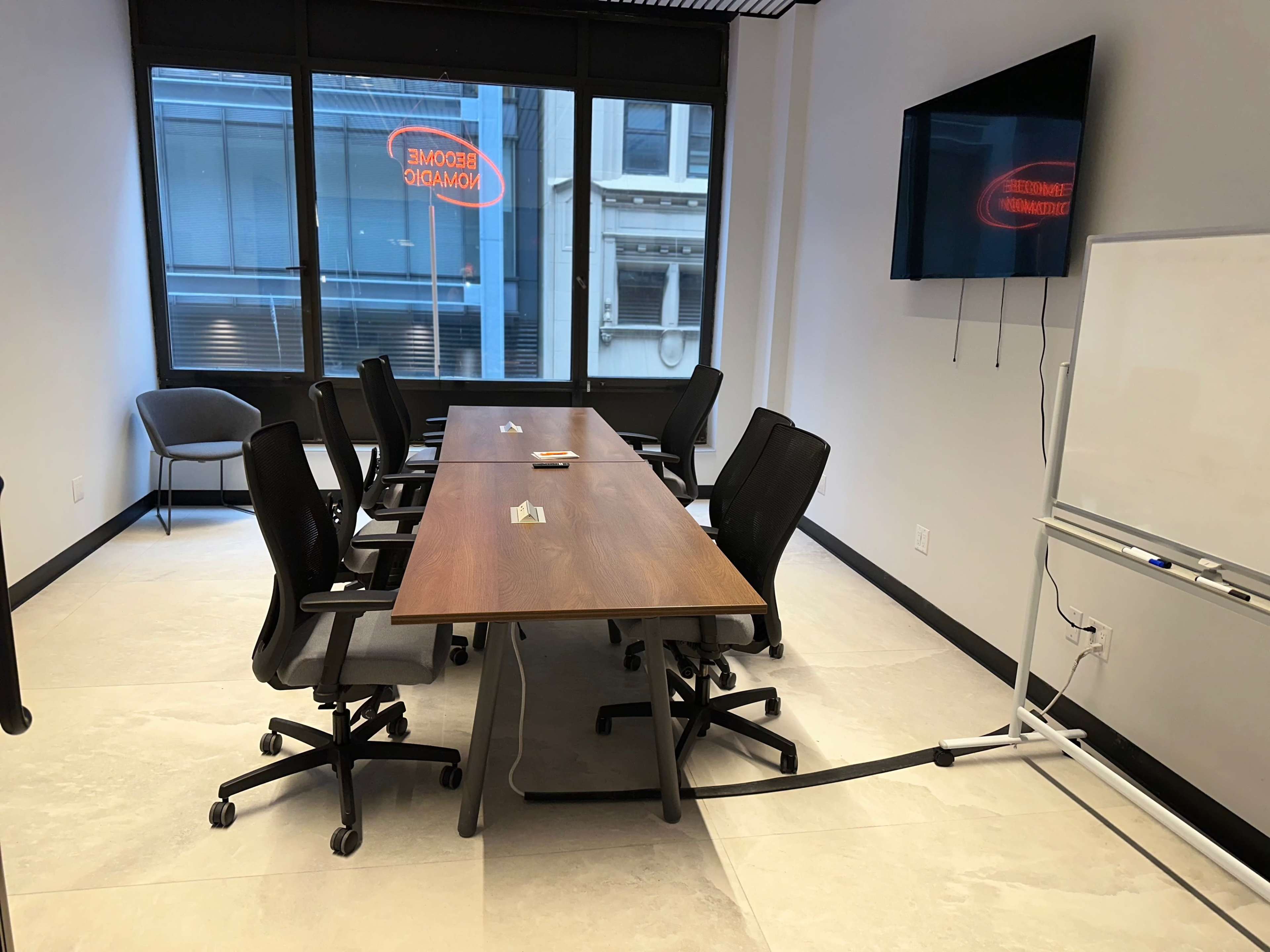 A conference room features a long wooden table surrounded by black rolling chairs, with a television mounted on the wall and large windows displaying a neon sign.