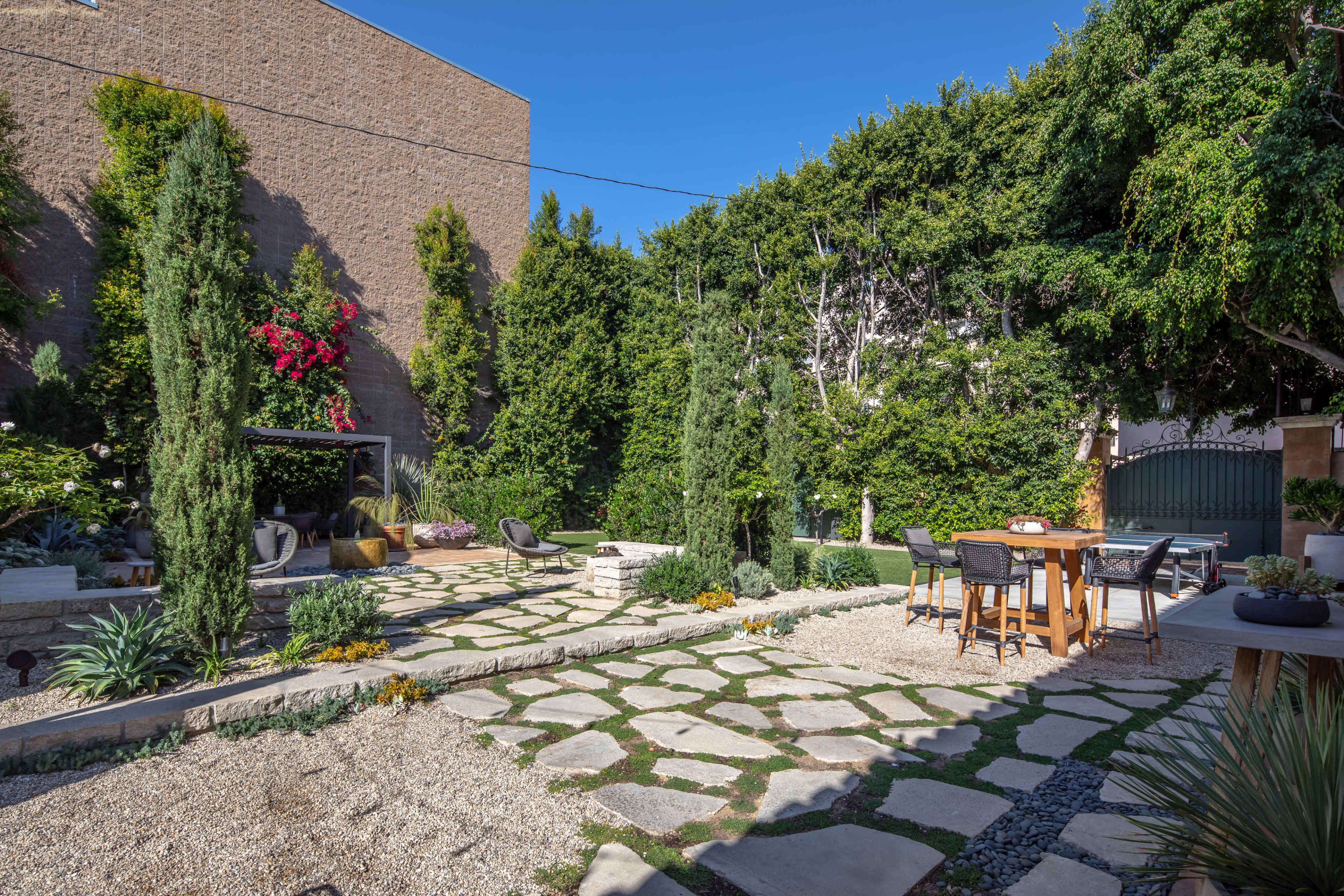 The image shows a landscaped backyard featuring stone pathways, greenery, and a dining area.