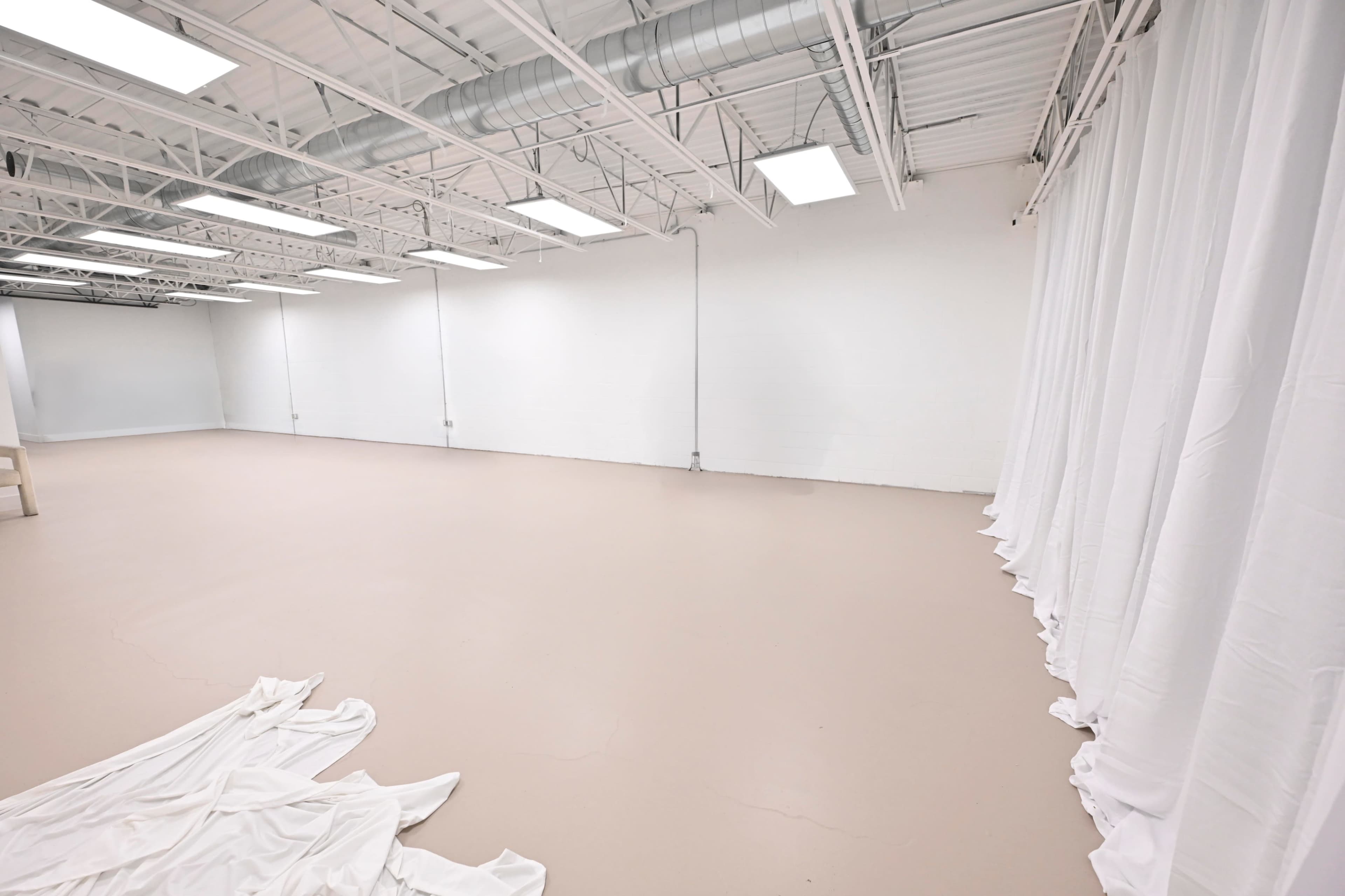 The image shows a spacious, empty room with a light-colored floor, white walls, and draped white fabric along one side.