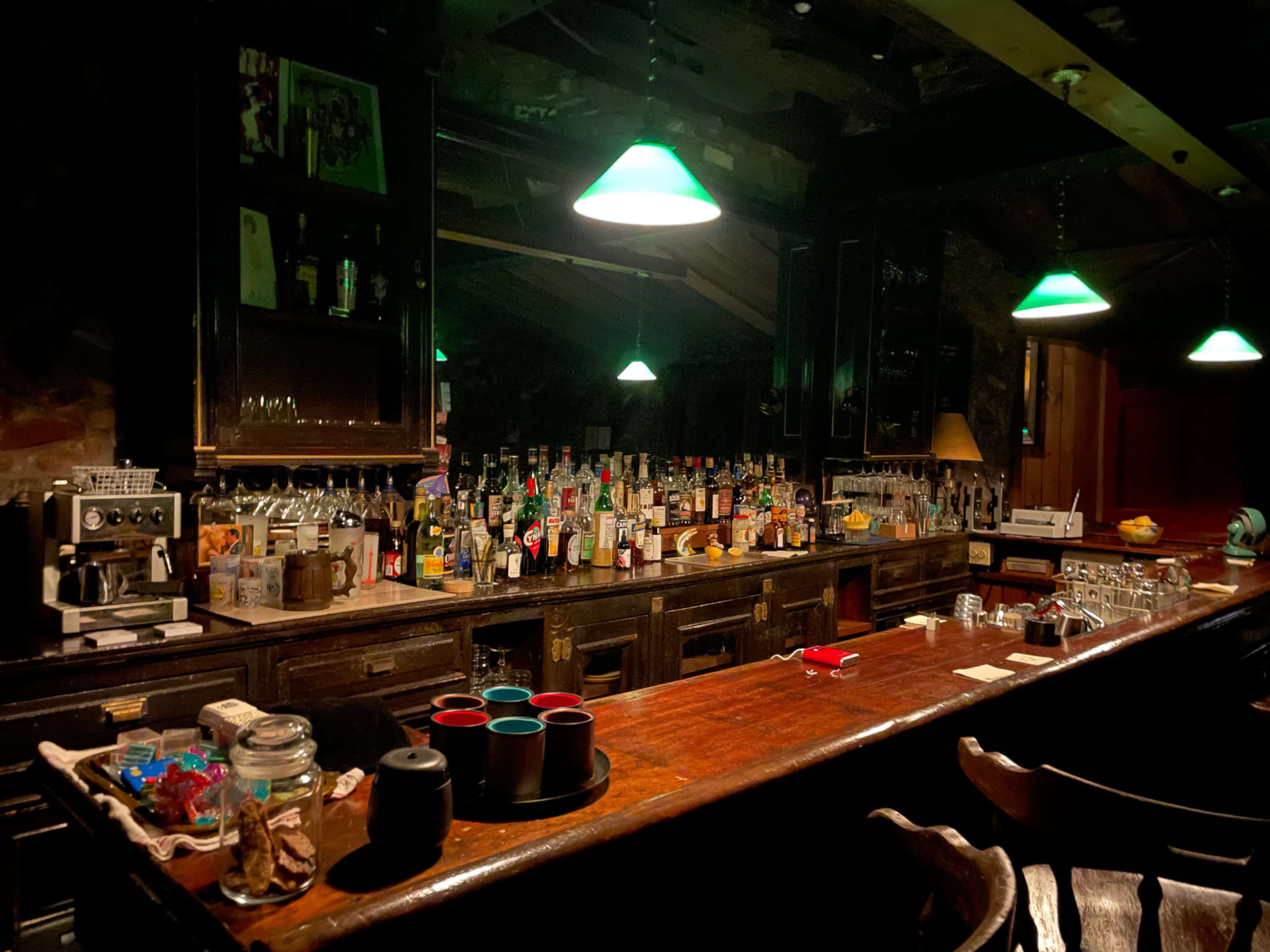 A dimly lit bar with a wooden counter, lined with numerous bottles and green pendant lights hanging overhead.