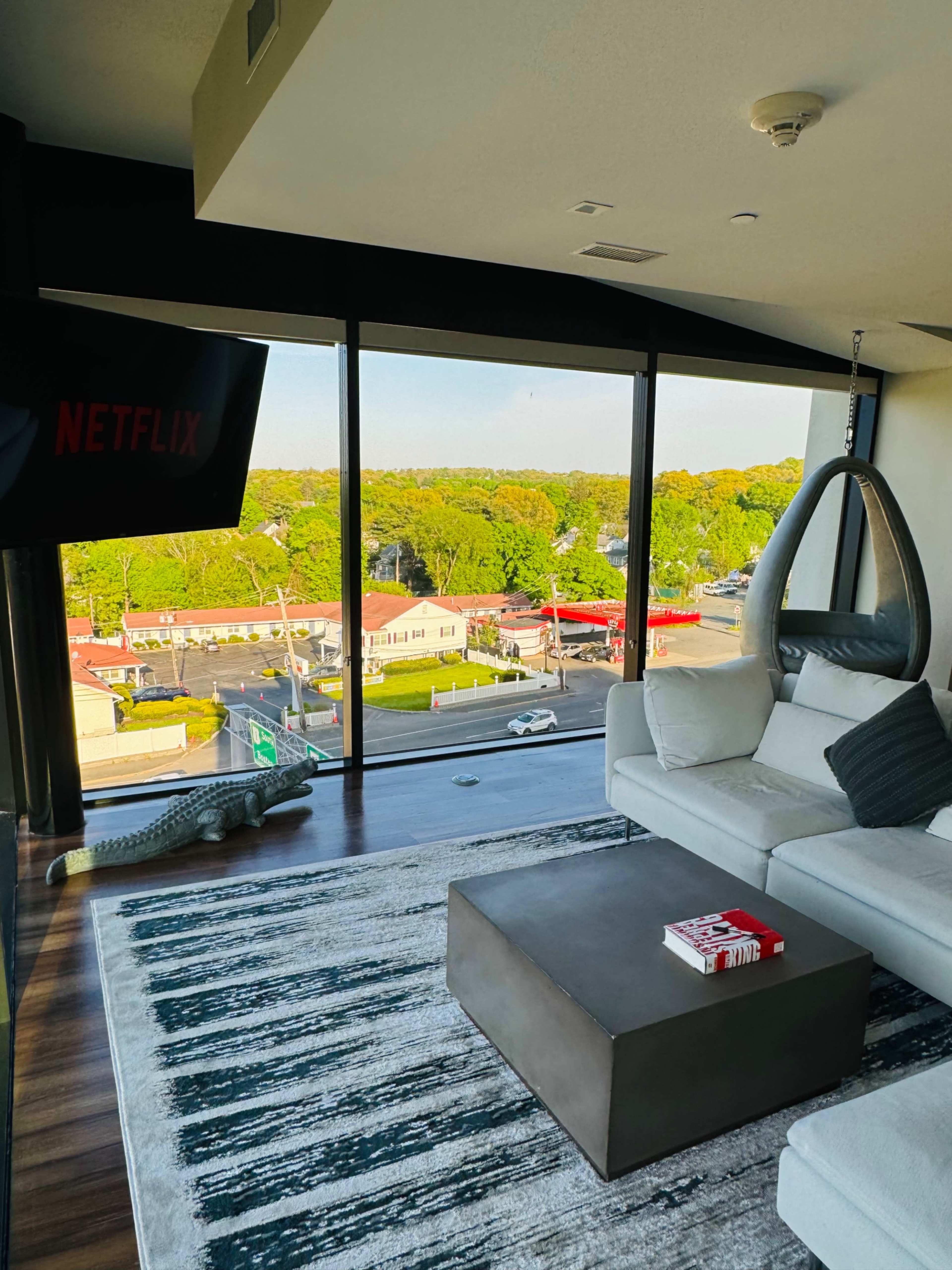 A modern living room features a large window overlooking a street lined with trees and buildings, with a hanging chair and a coffee table in the foreground.