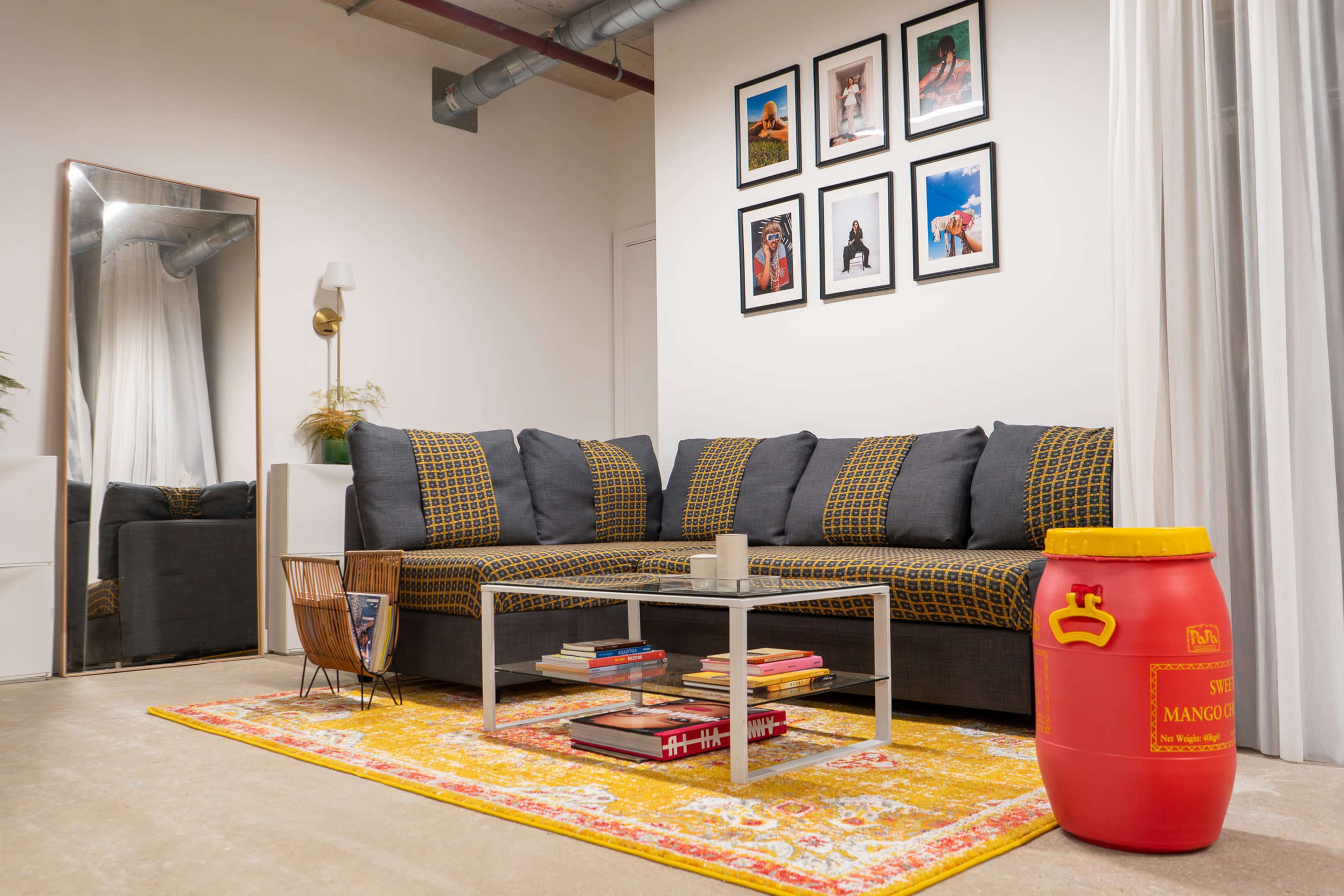 The image shows a modern living room featuring a gray sectional sofa, a glass coffee table with stacked books, a decorative yellow rug, and a bright yellow barrel-style storage container.
