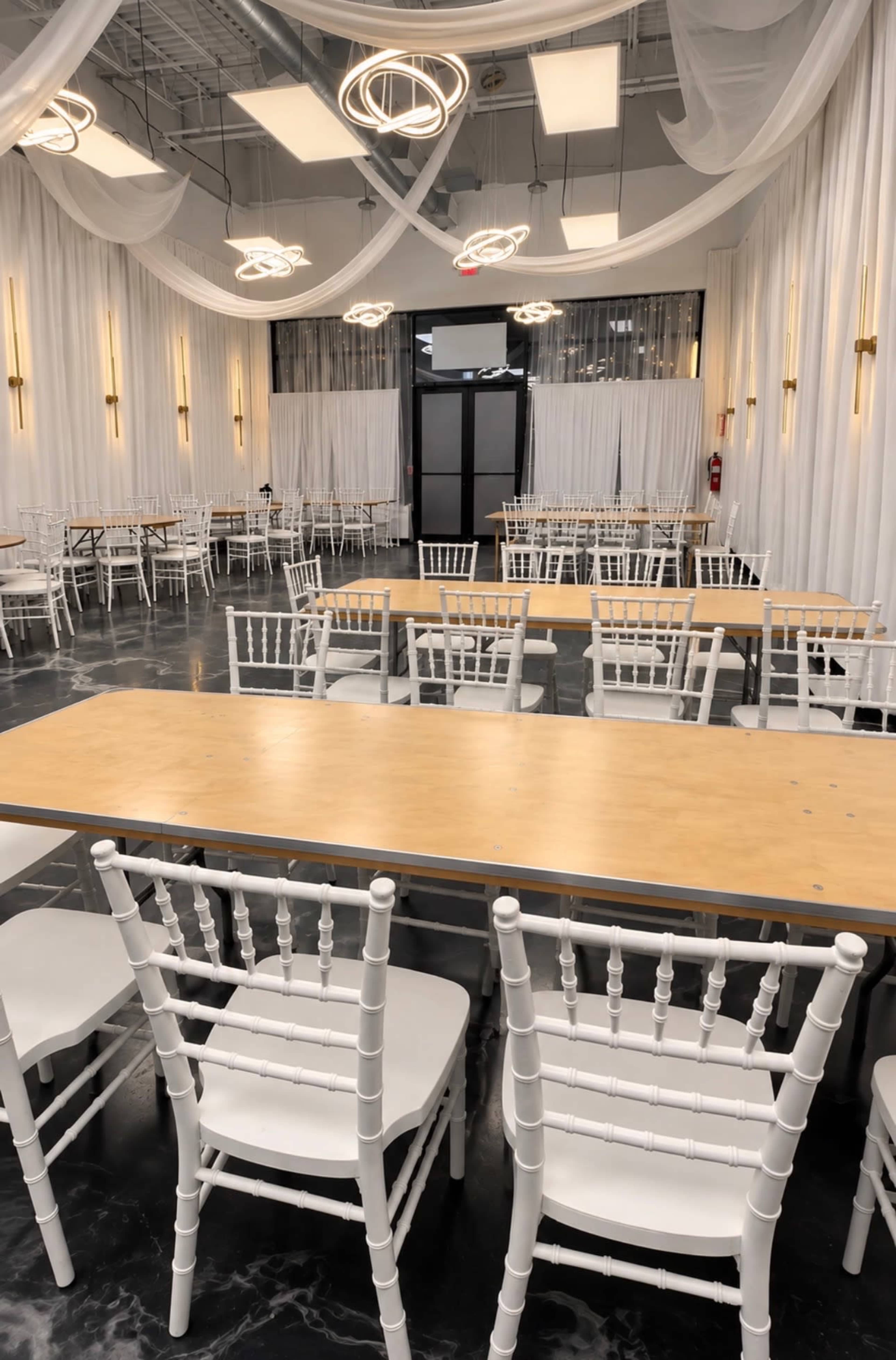The image shows a spacious event room with white draped walls, multiple tables arranged in rows, and elegant light fixtures hanging from the ceiling.