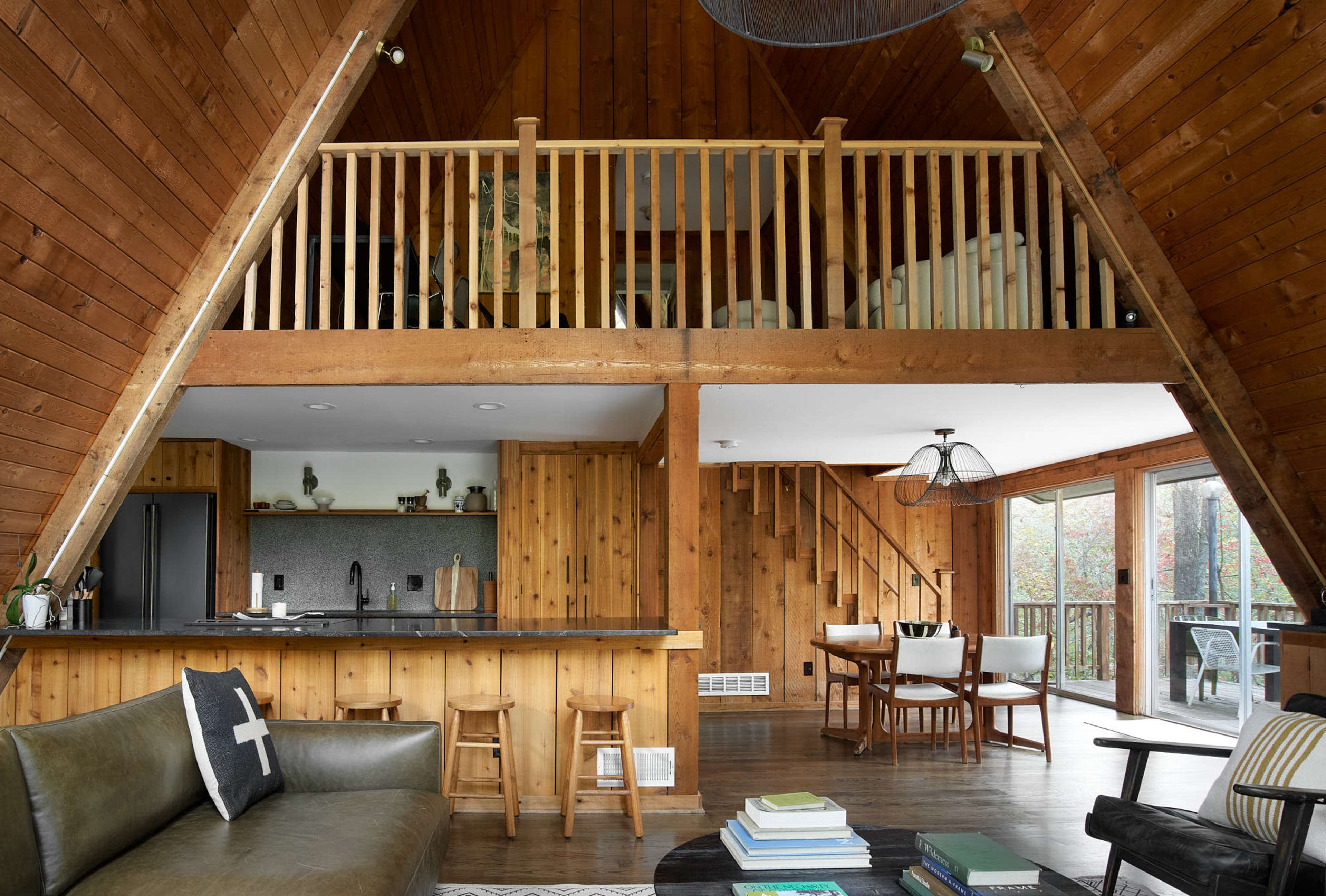 An interior of a wooden A-frame cabin featuring a kitchen, dining area, and a lofted sleeping space.
