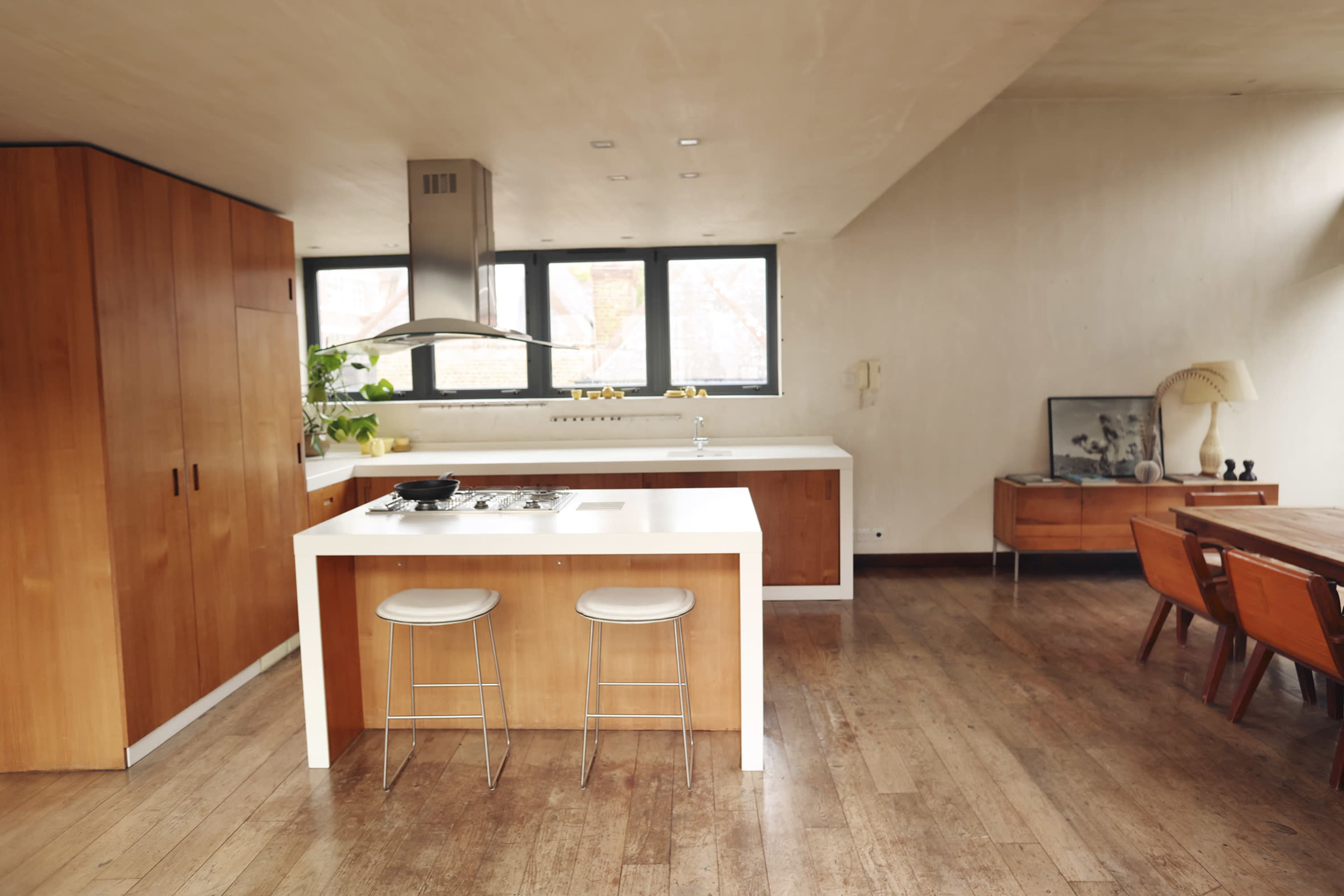 The image shows a modern kitchen with wooden cabinetry, a central island with a stove, and large windows providing natural light.