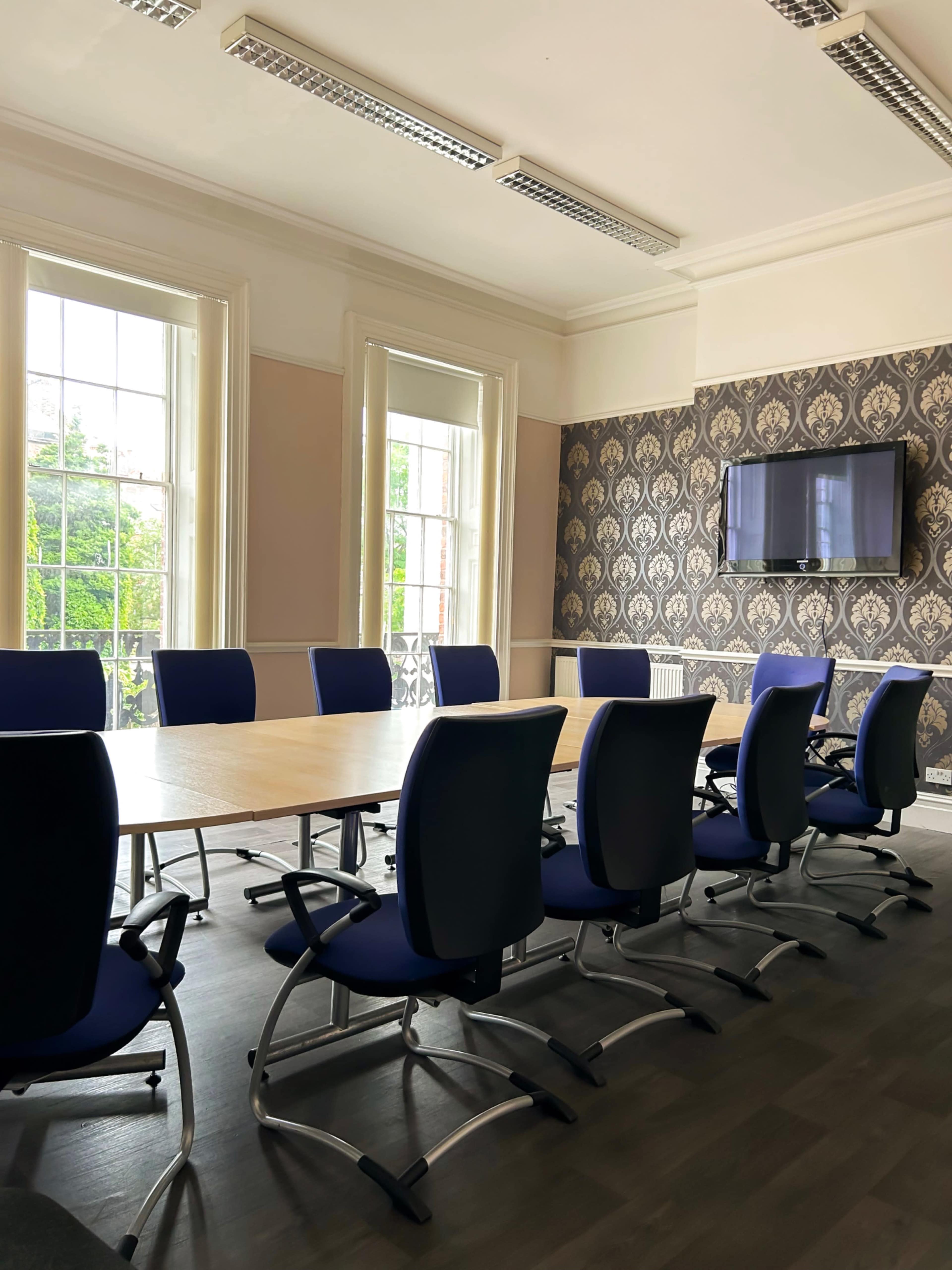 The image shows a conference room with a long wooden table surrounded by blue office chairs, large windows letting in natural light, and a wall-mounted television.