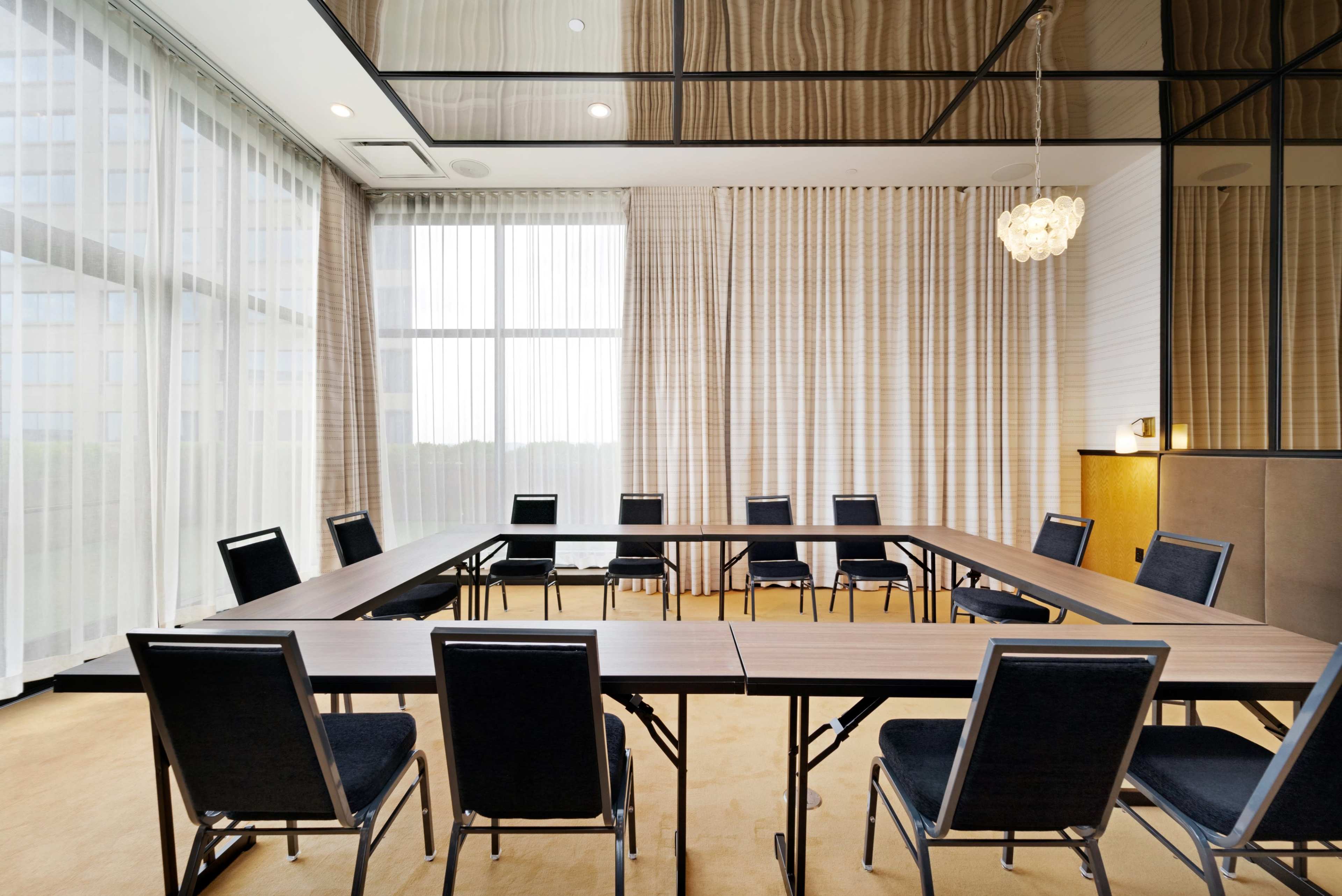 A conference room features a rectangular arrangement of wooden tables surrounded by black chairs, with large windows allowing natural light to enter.