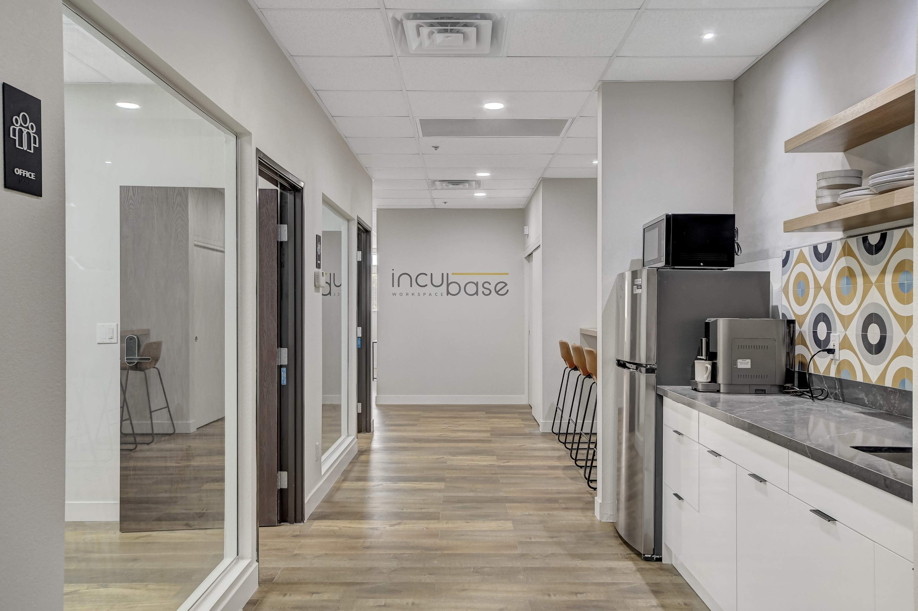 A modern office space with a kitchenette on one side and two glass-walled offices on the other, featuring wood flooring and contemporary design elements.