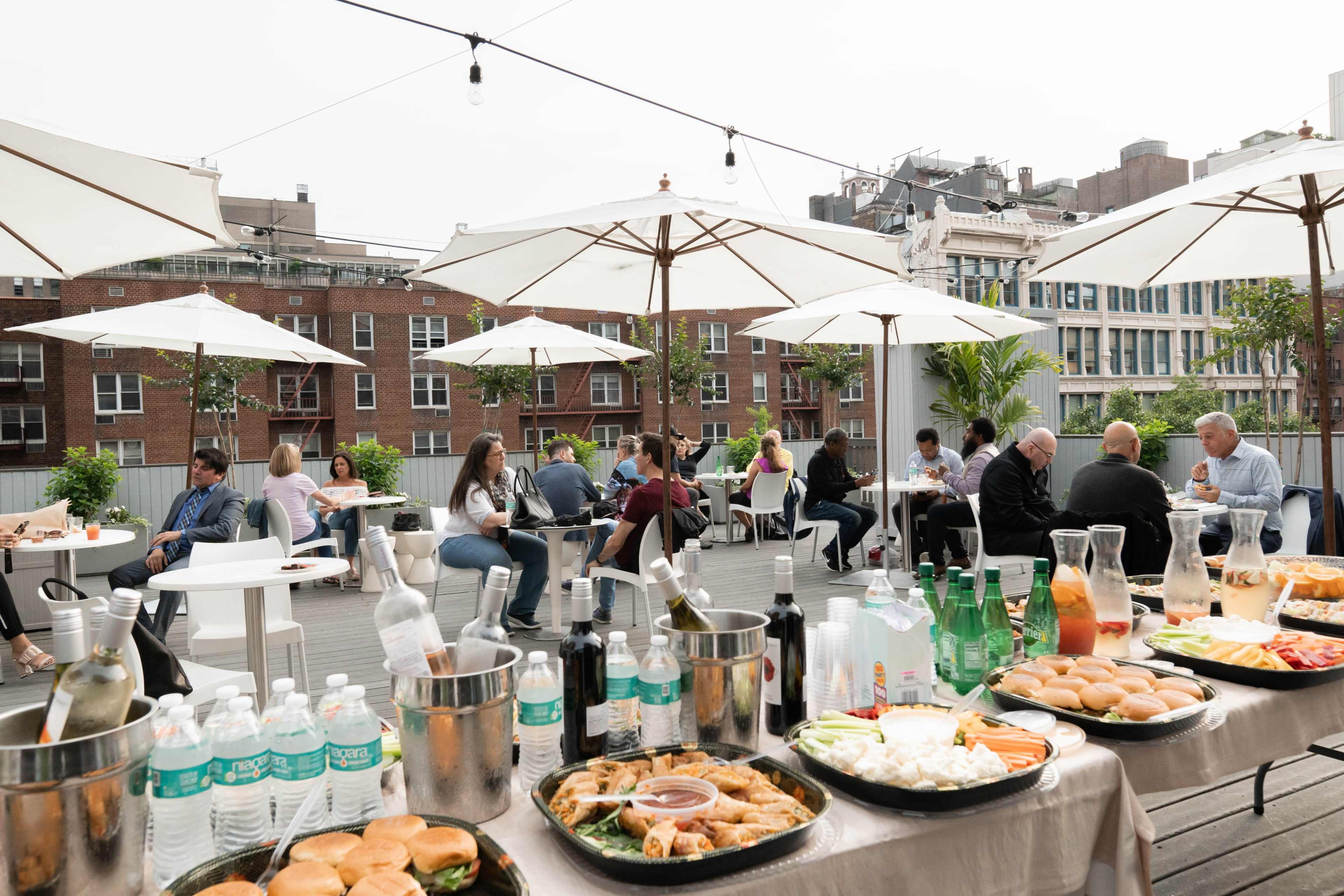 A rooftop gathering features tables with food and drinks, umbrellas for shade, and people socializing in a city setting.