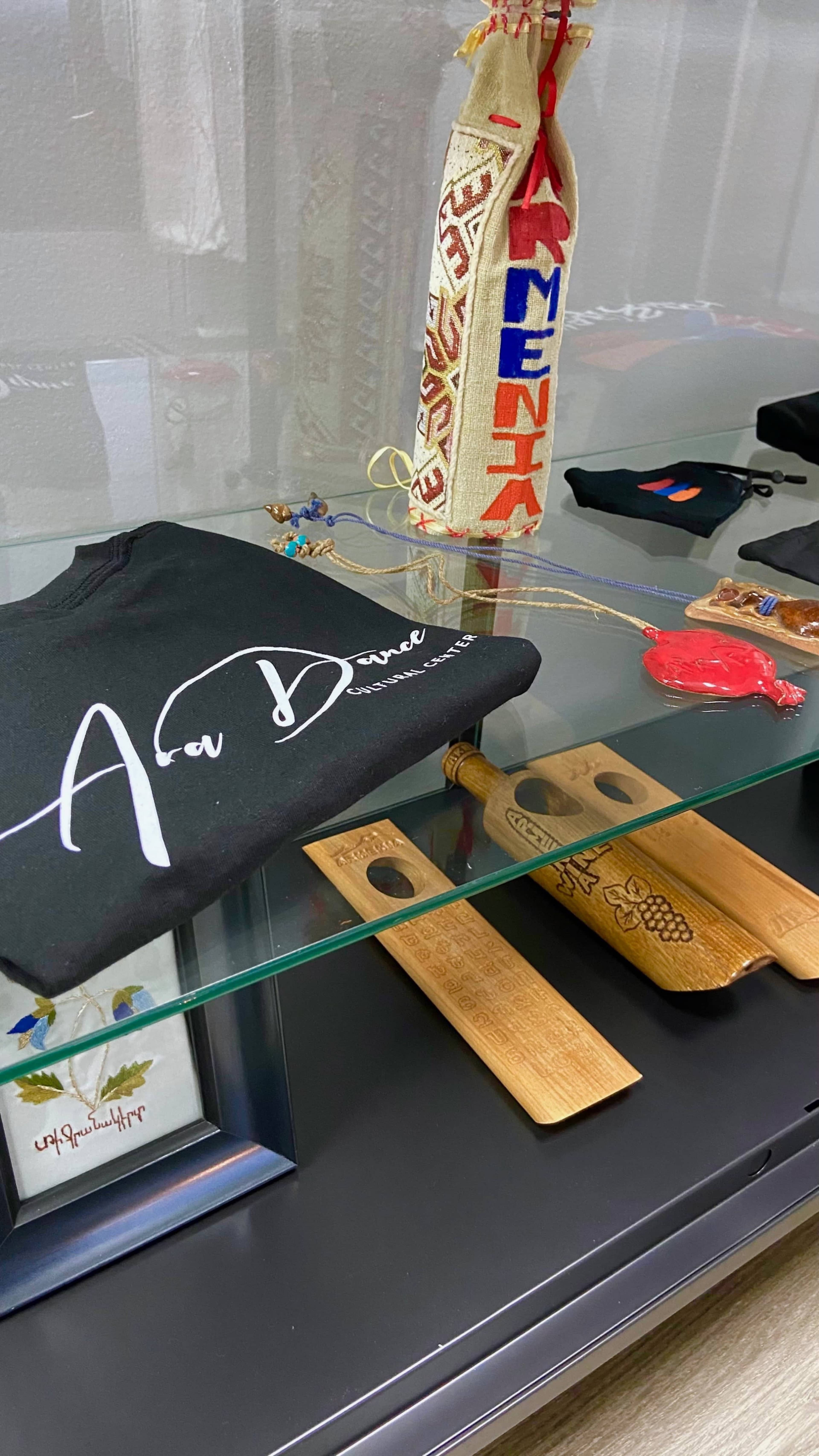 A display of various items including a black t-shirt with "Ava Dance" printed on it, wooden utensils, and a decorative bag, all arranged on a glass shelf.